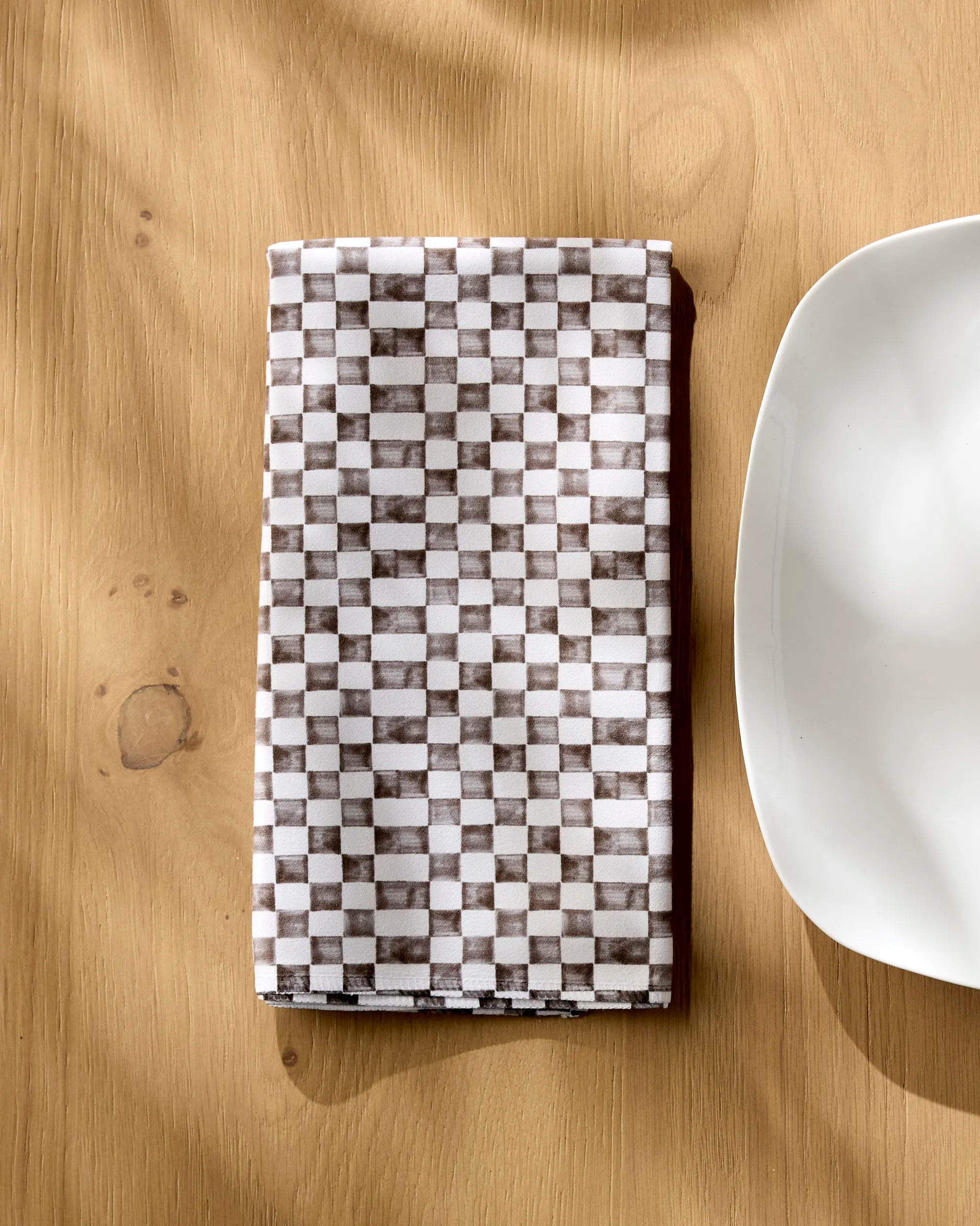 The Geometry Tavern napkin, featuring gray and white squares, rests folded on a light wooden table by a white, round-cornered plate as sunlight casts soft shadows over the scene.
