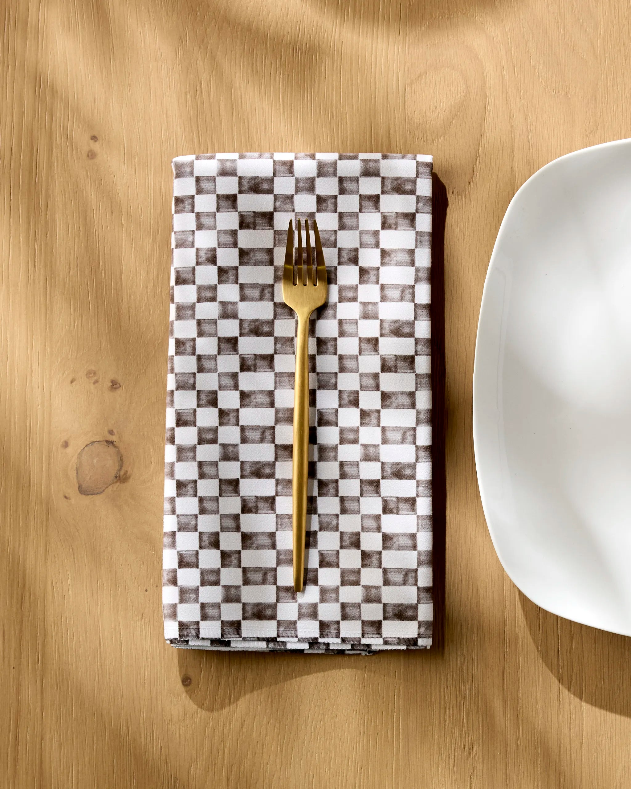 A gold fork rests on a folded Tavern napkin by Geometry, featuring a brown and white checkered pattern, placed on a light wooden table beside the edge of a white plate.
