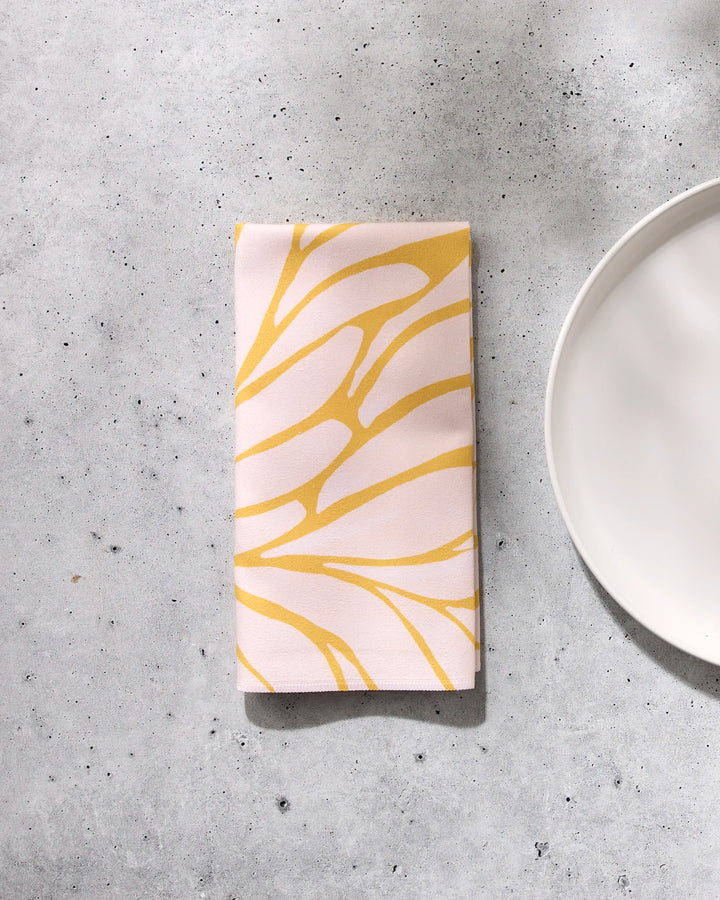The GEOMETRY "Tenderhearted" napkin features a yellow abstract leaf pattern on a light background, folded and placed on a gray speckled surface beside the edge of a white plate.