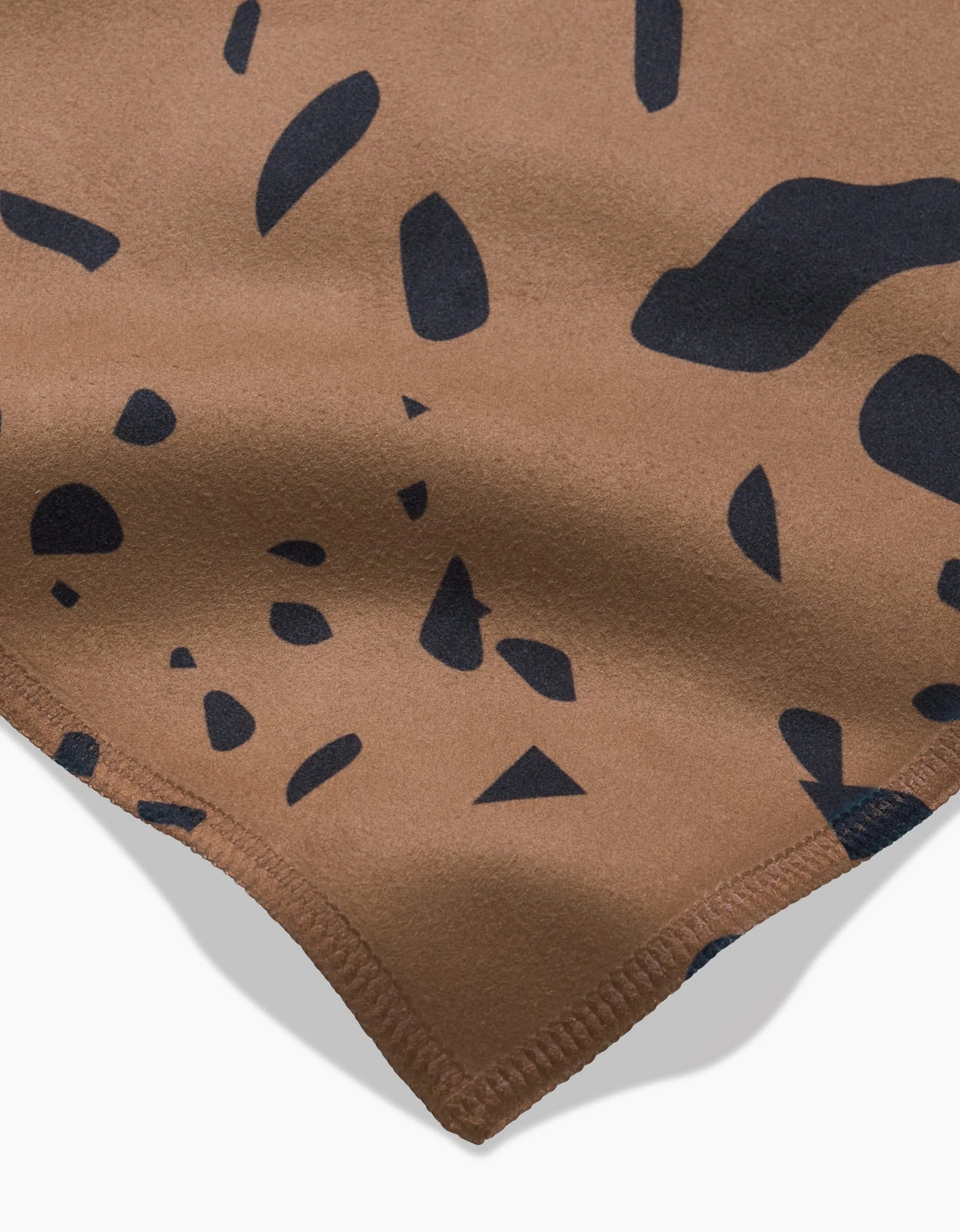 Close-up of GEOMETRYs Terrazo: brown fabric with black abstract spots and a stitched edge, inspired by animal print—ideal for recycled dinner napkins or as part of an absorbent cloth napkin set.