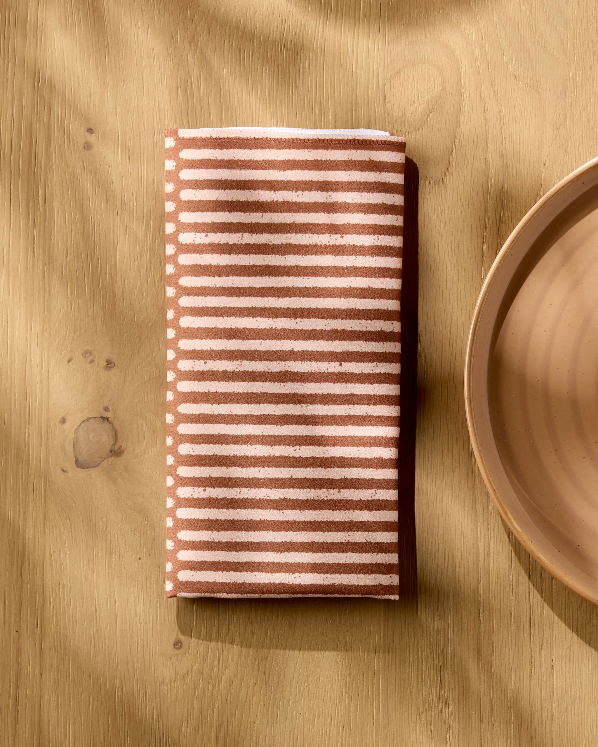 A Rosa Kichline Textured Stripe reusable napkin with cream and brown stripes rests on a light wooden surface beside a beige ceramic plate, sunlight casting soft shadows across this eco-friendly dinner napkin set.