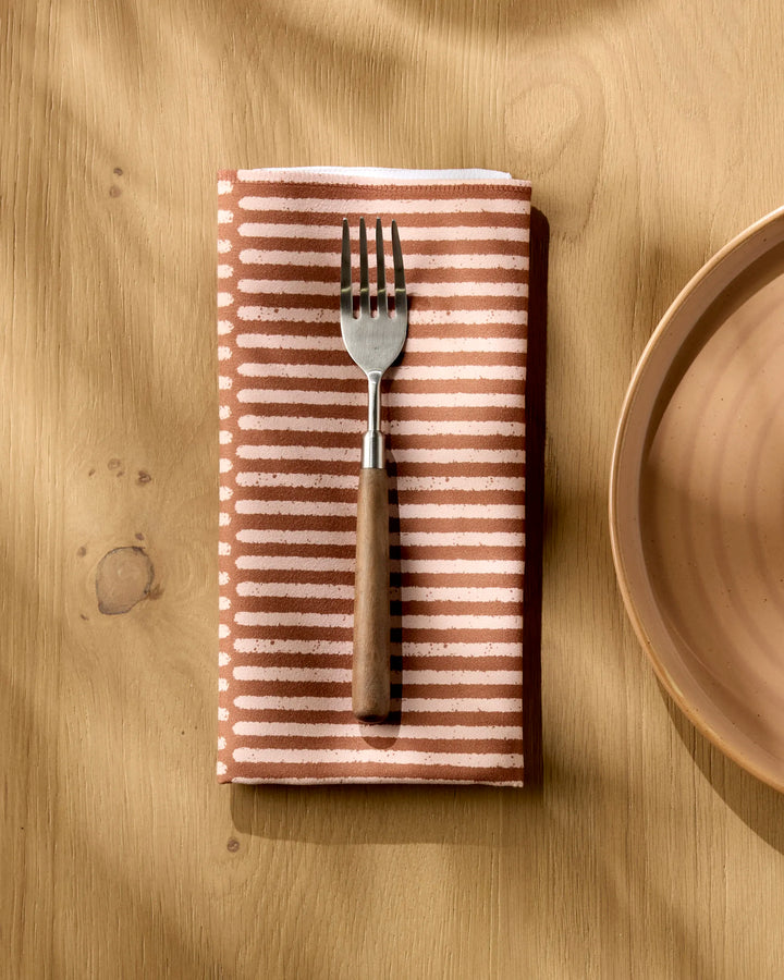 A fork with a wooden handle rests on Rosa Kichline’s Textured Stripe reusable napkin beside a ceramic plate on a wooden table, as sunlight casts soft shadows across the scene.