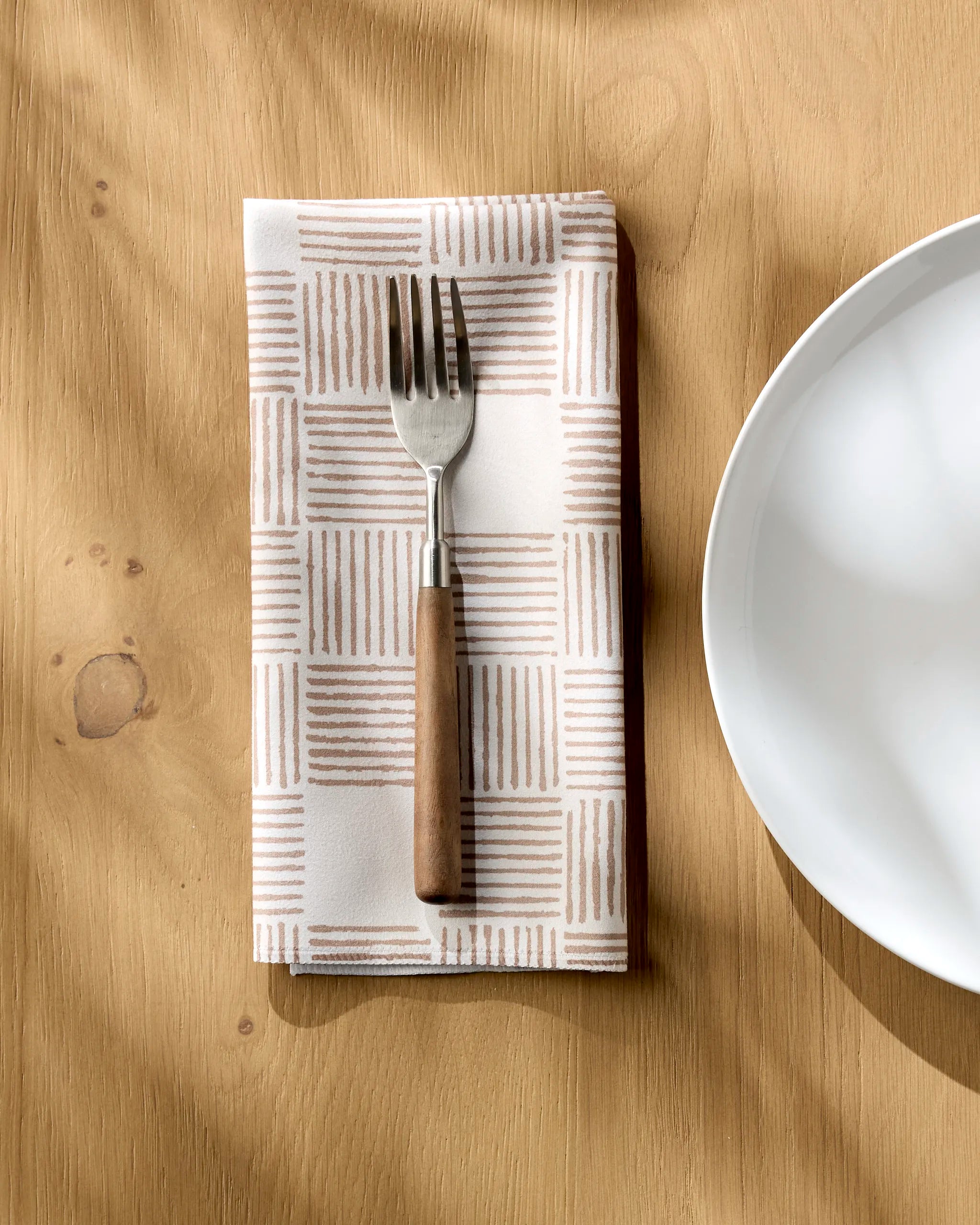 The Thatcher fork by Studio Jeannot, featuring a wooden handle, lies on an eco-friendly napkin beside a white plate on a light wooden table, sunlight casting gentle shadows.