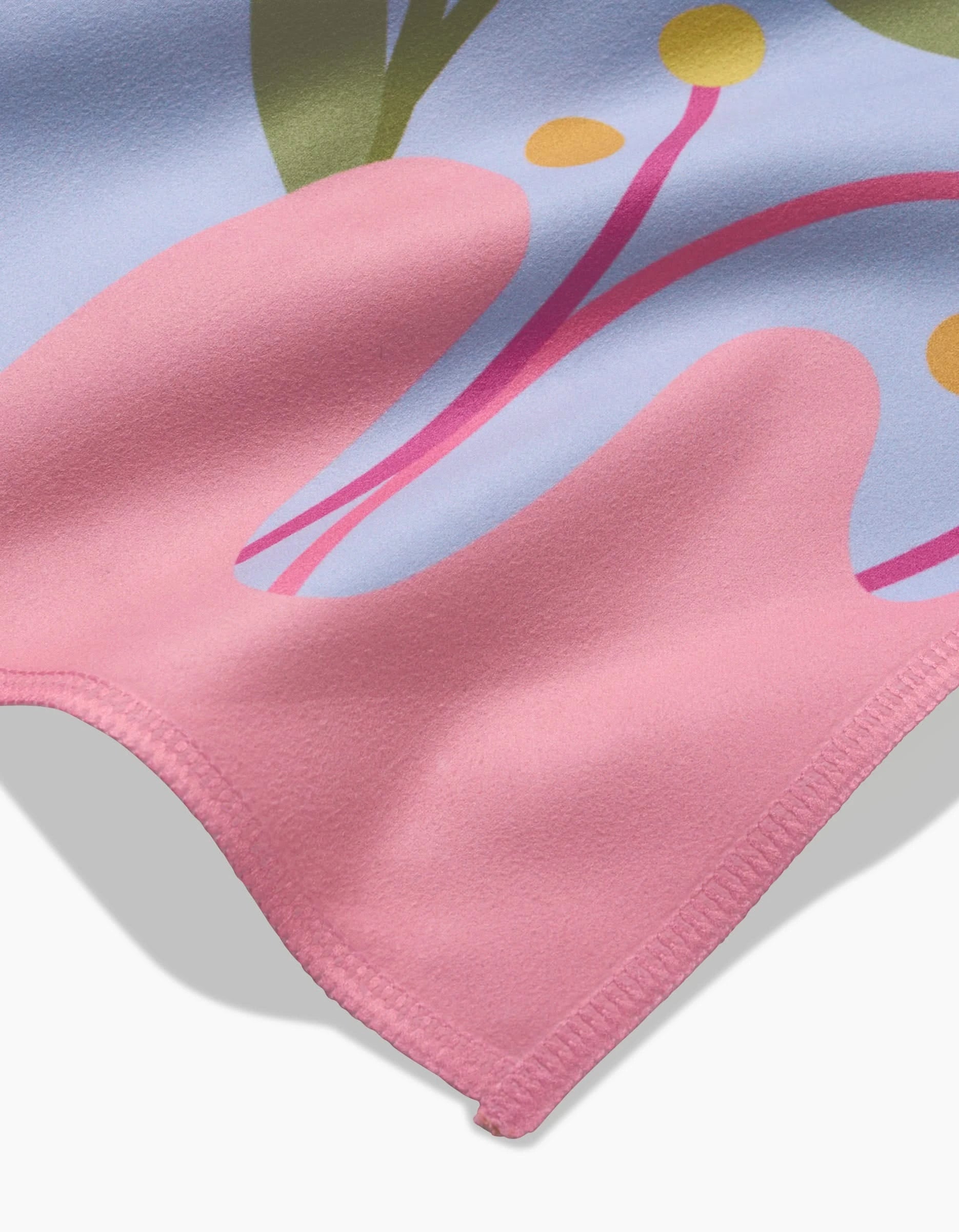 The GEOMETRY Thoughtful blanket features soft pink Geosuede fabric with a vibrant abstract pattern—curved green, purple, and yellow shapes. Stitched edges and recycled materials offer eco-friendly comfort and style, shown here in a close-up view.