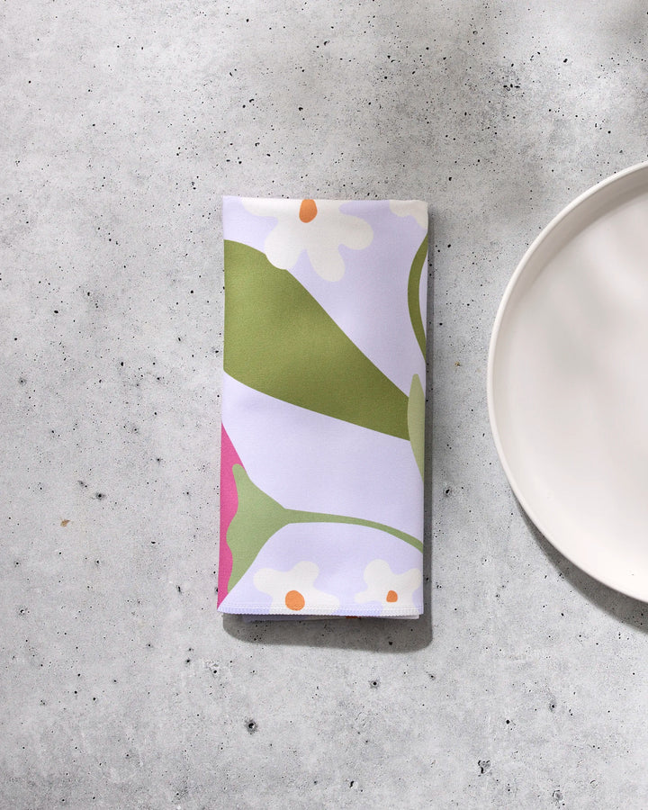 The GEOMETRY Thoughtful napkin, featuring a vibrant green, white, orange, and magenta floral design, is folded on a light gray textured surface beside a white round plate.