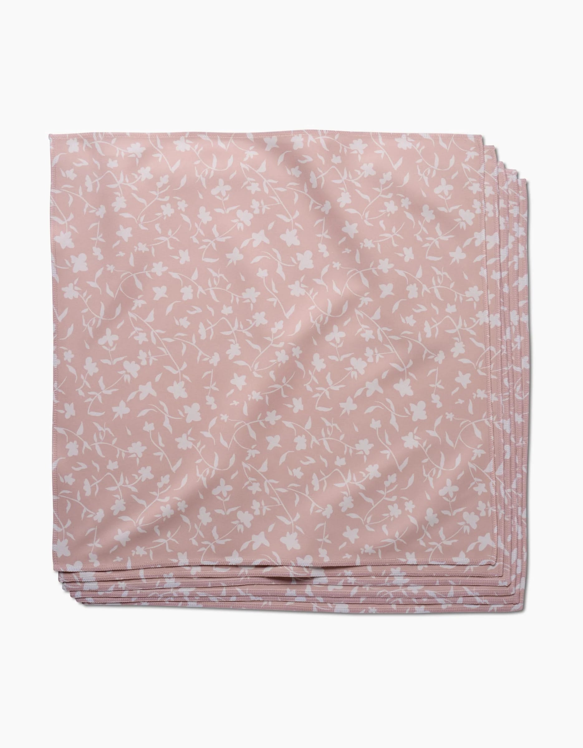 A neatly folded, pale pink Tiny Floral dinner napkin by Aubrey Fairchild, made from soft Geosuede fabric with a white floral pattern, is stacked with identical napkins on a white background.