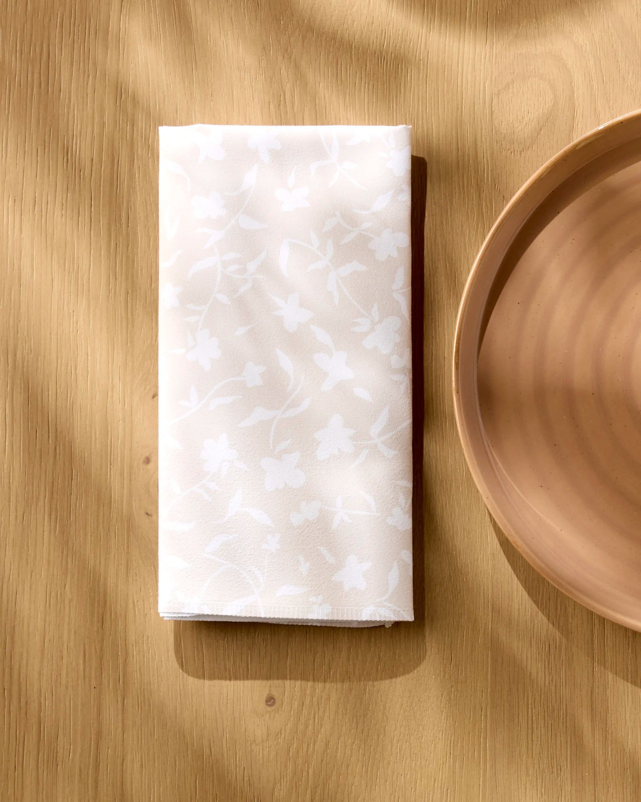 The Aubrey Fairchild Tiny Floral white reusable dinner napkin, featuring a subtle floral pattern, is folded on a light wooden table beside a round beige plate, illuminated by soft natural light and gentle shadows.