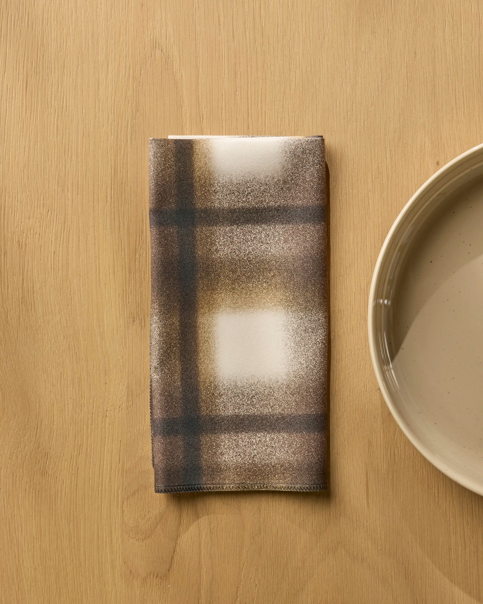 A Geometry "Toasted" brown and white plaid-patterned absorbent cloth napkin sits folded on a light wooden table beside a beige ceramic plate.