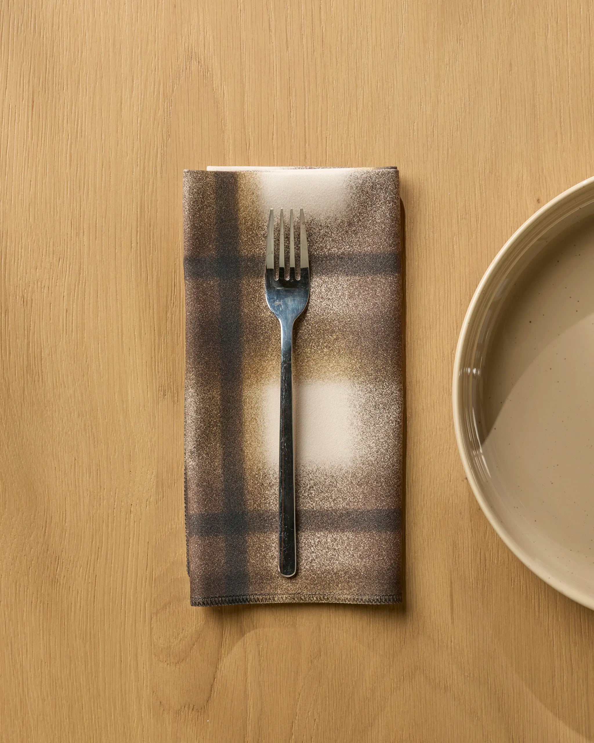 A metal fork rests on a Geometry Toasted brown and beige plaid dinner napkin set, neatly folded on a light wooden table, with part of a beige ceramic plate visible on the right.