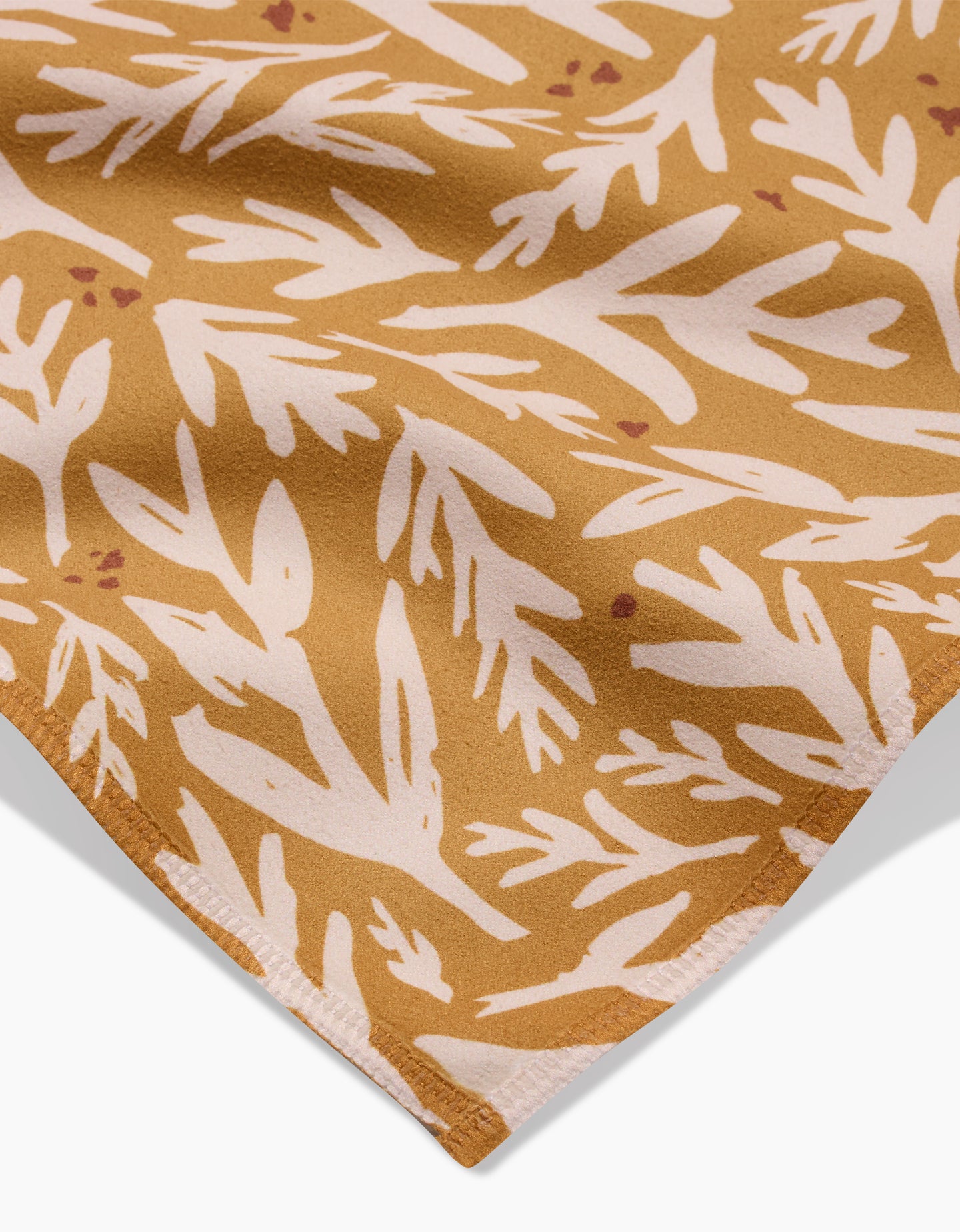 A close-up of Twig + Sparks Trail Dusting Gold napkin, a brown absorbent cloth with a white abstract leafy pattern and neatly stitched edge.
