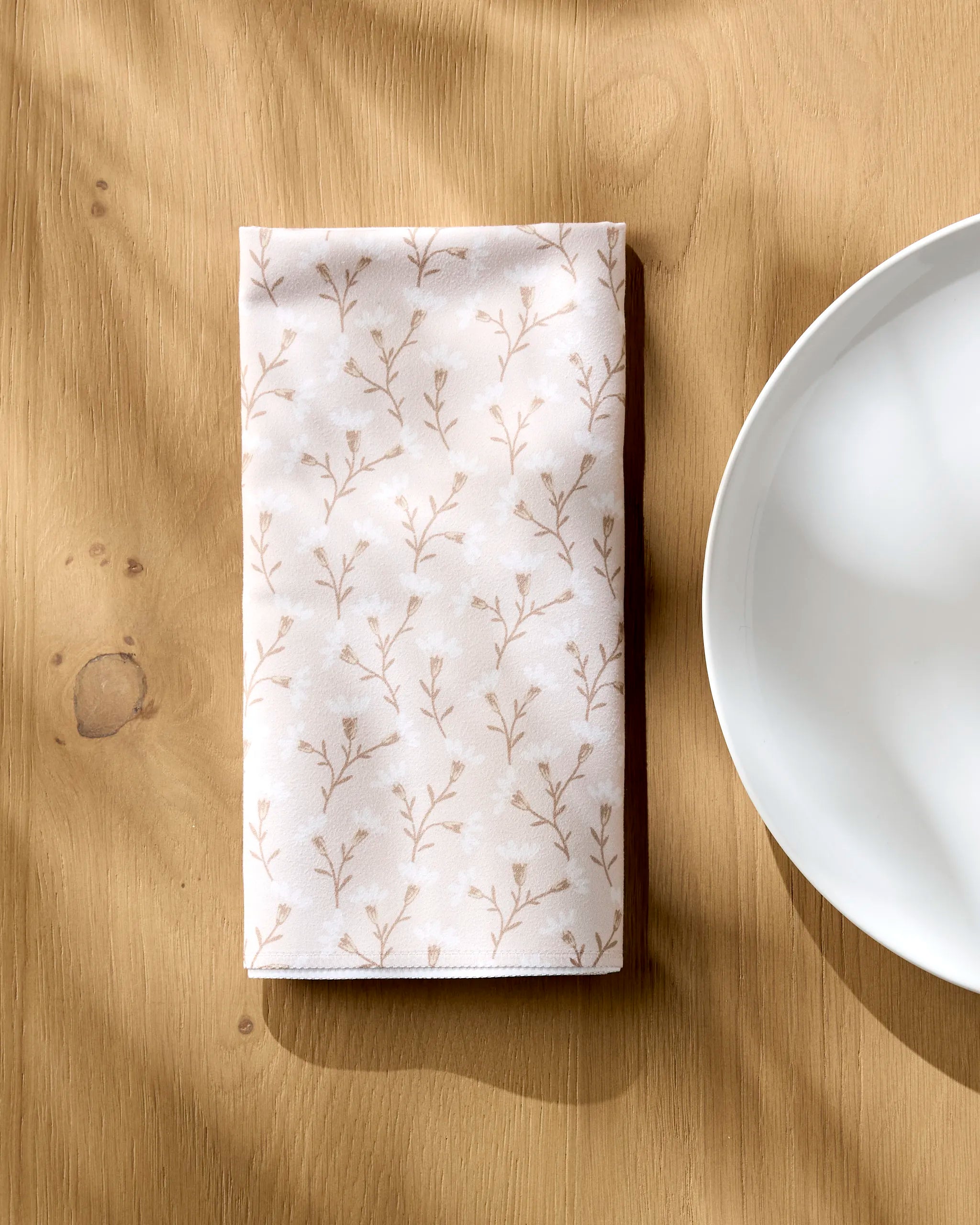A folded Trailing Flowers in Pink reusable dinner napkin by Teri the Butter, featuring a delicate floral pattern, rests on a wooden table beside a white plate, with sunlight casting gentle shadows over the scene.
