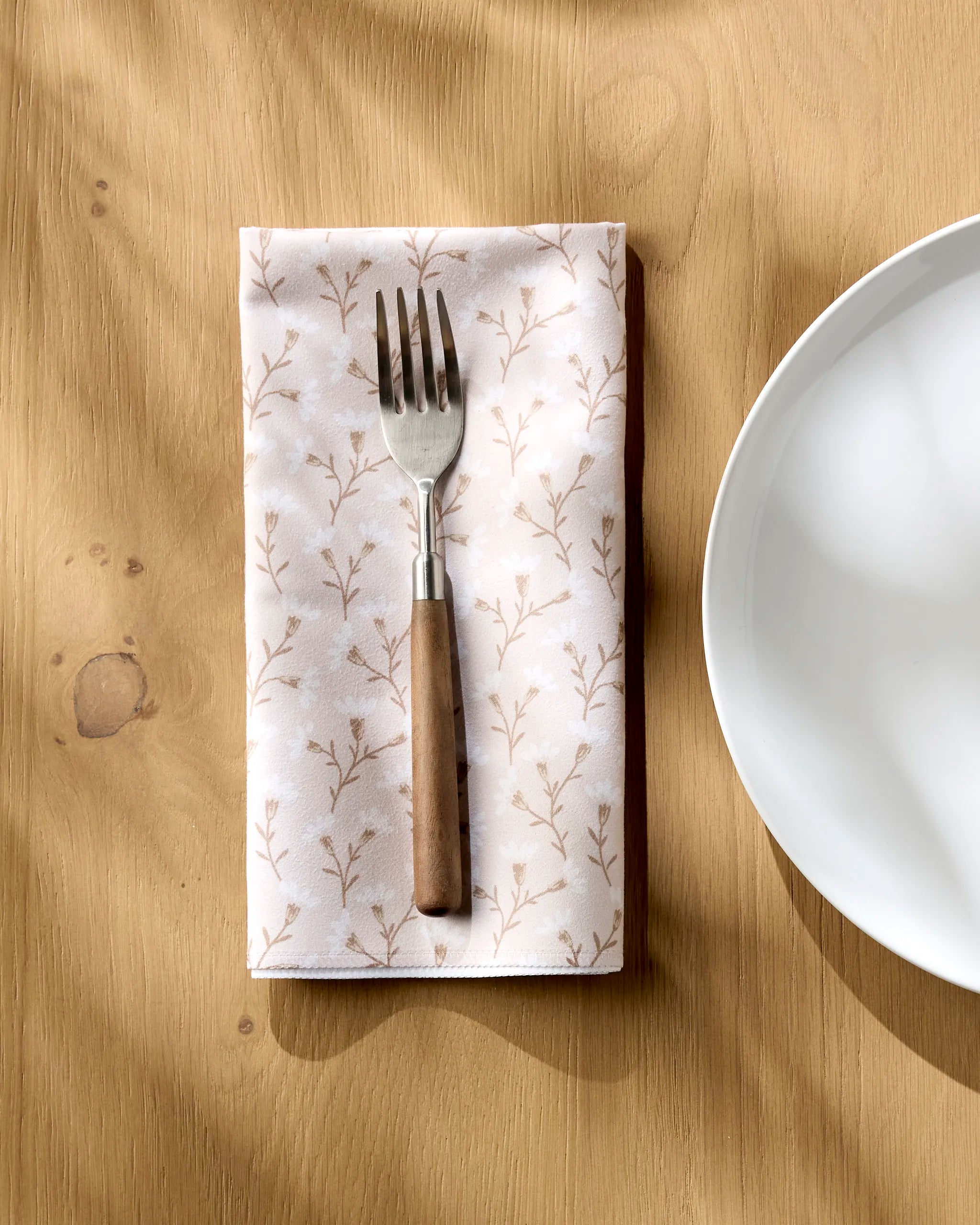 A fork sits on the Teri the Butter "Trailing Flowers in Pink" reusable napkin, placed on a wooden table beside an empty white plate, with sunlight casting gentle shadows over the scene.