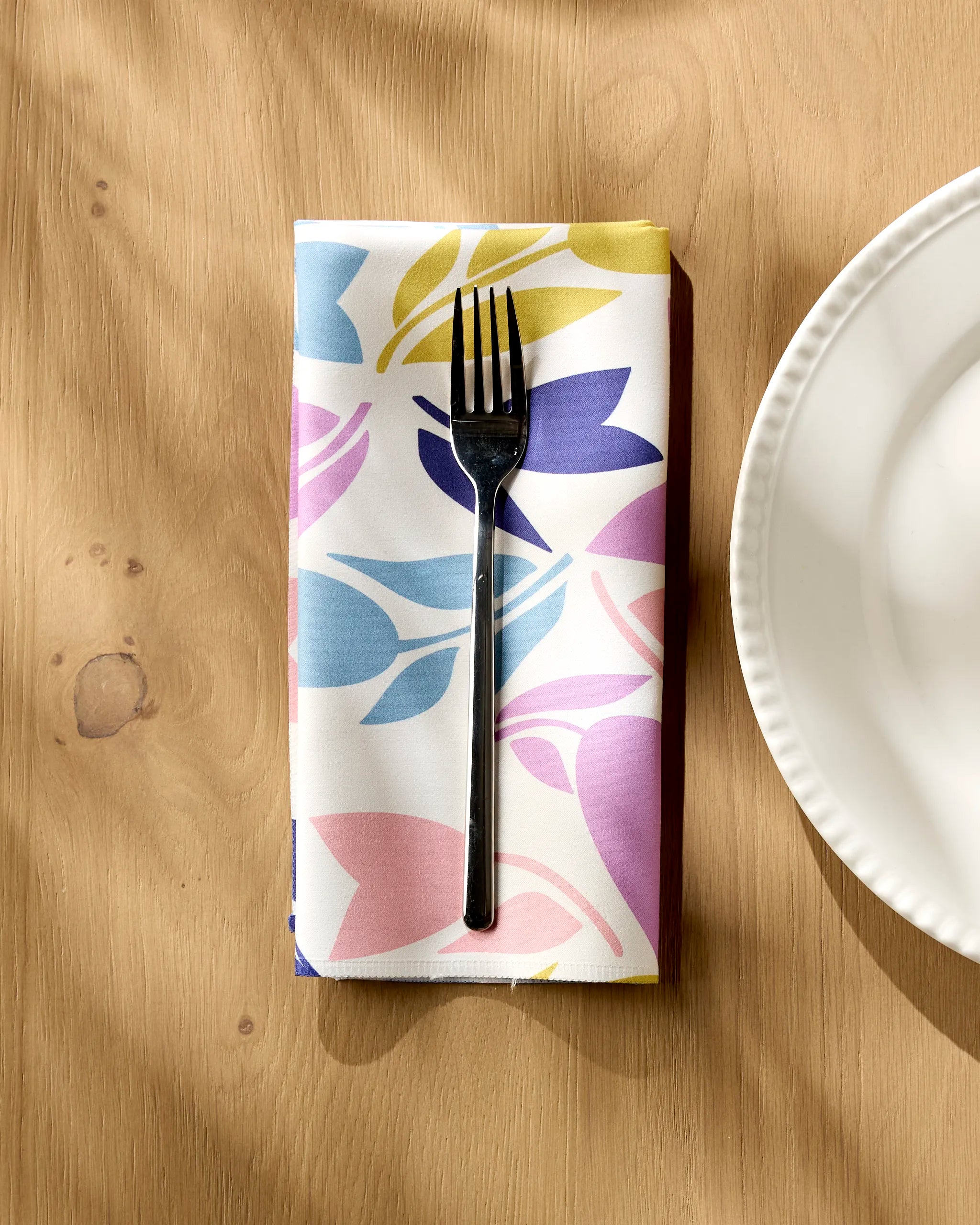 A fork rests on Lizzie Clark's Tulip Tumble colorful reusable cloth napkin, neatly folded on a light wooden table next to a white plate, as sunlight casts soft shadows across the scene.