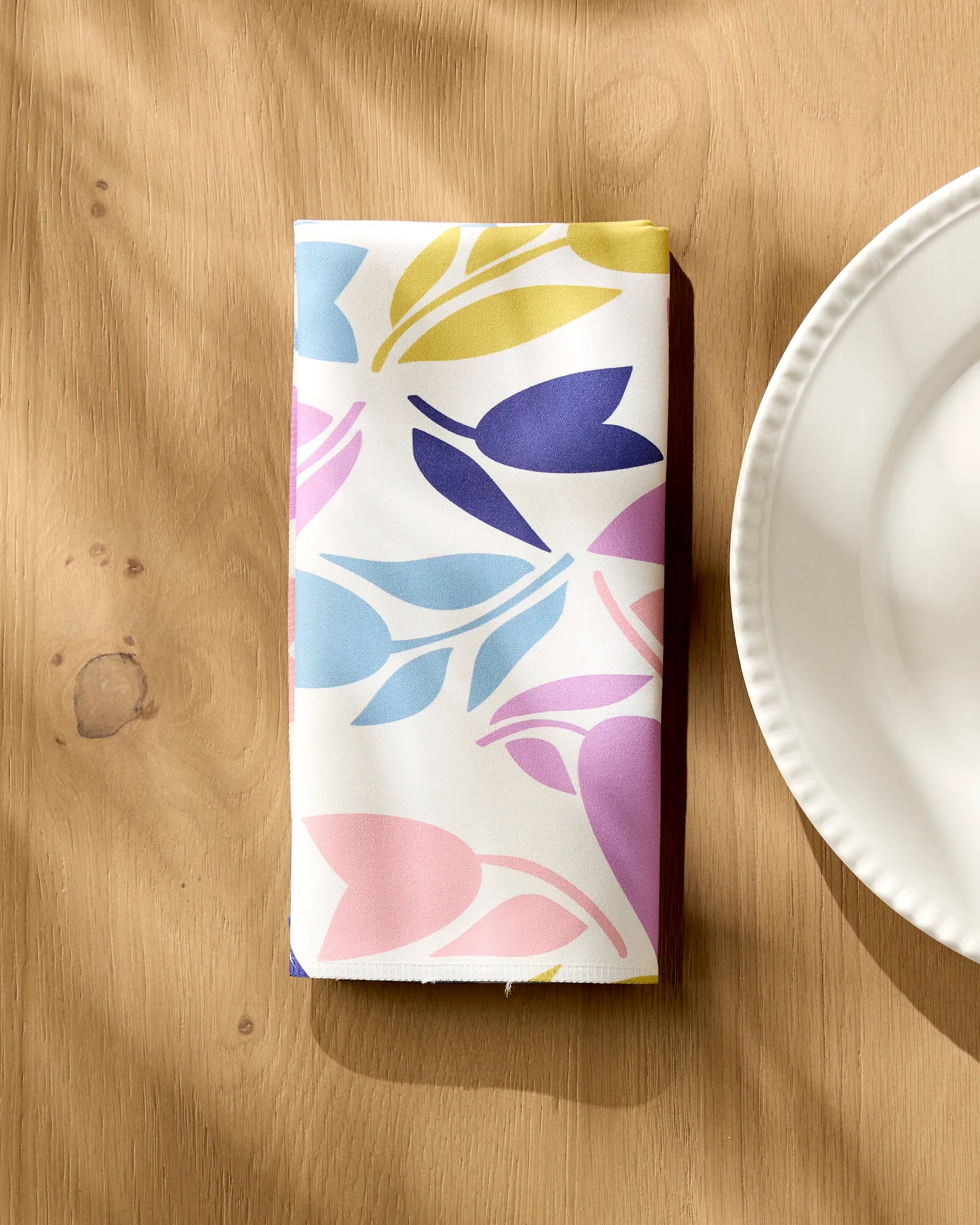 A folded Lizzie Clark "Tulip Tumble" reusable cloth napkin with a colorful floral pattern rests on a wooden table beside a white plate, sunlight casting soft shadows across the scene.