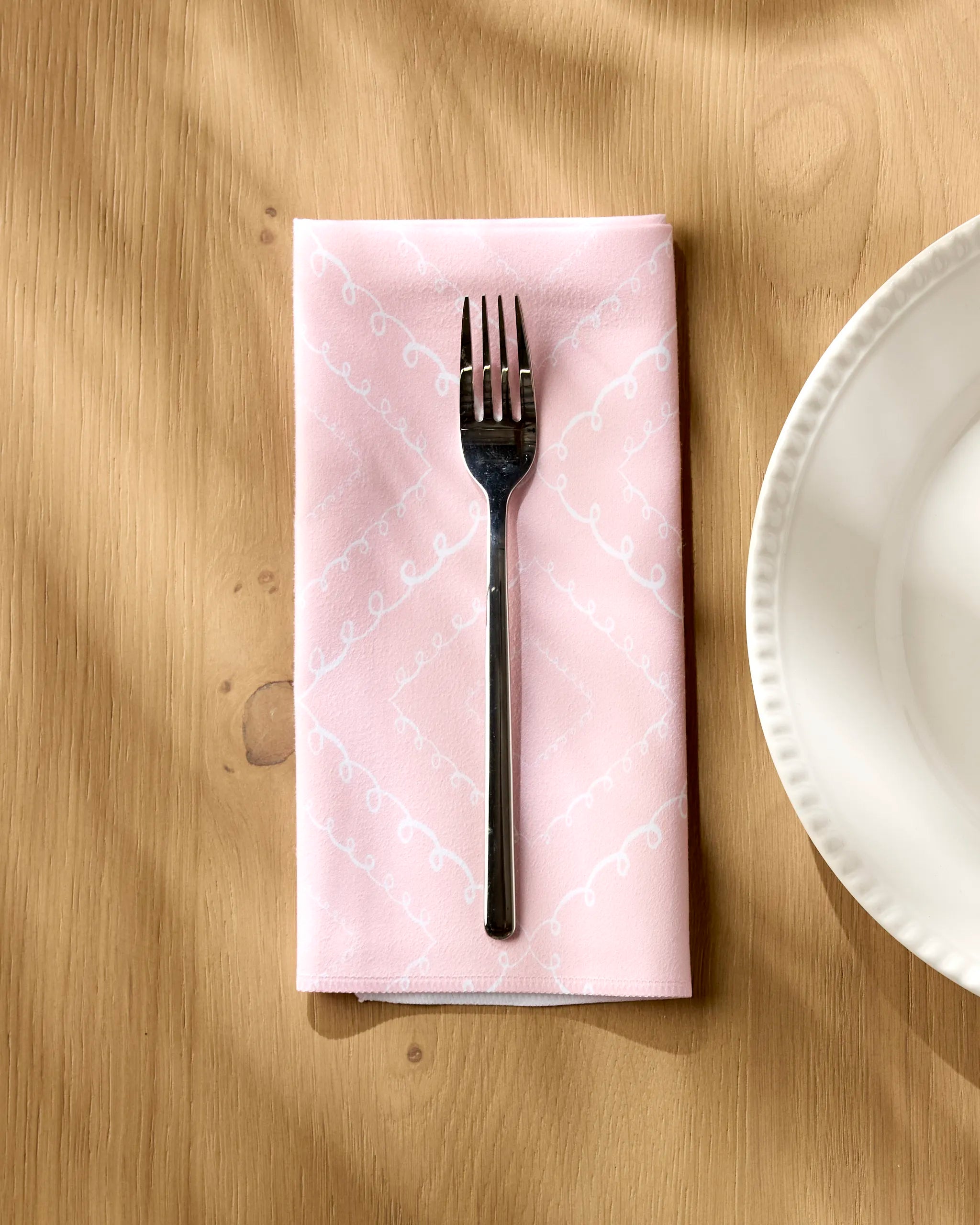 A silver fork rests on Elena Fay's Twirls pink recycled dinner napkin with a white pattern, atop a wooden table beside a scalloped-edge white plate.