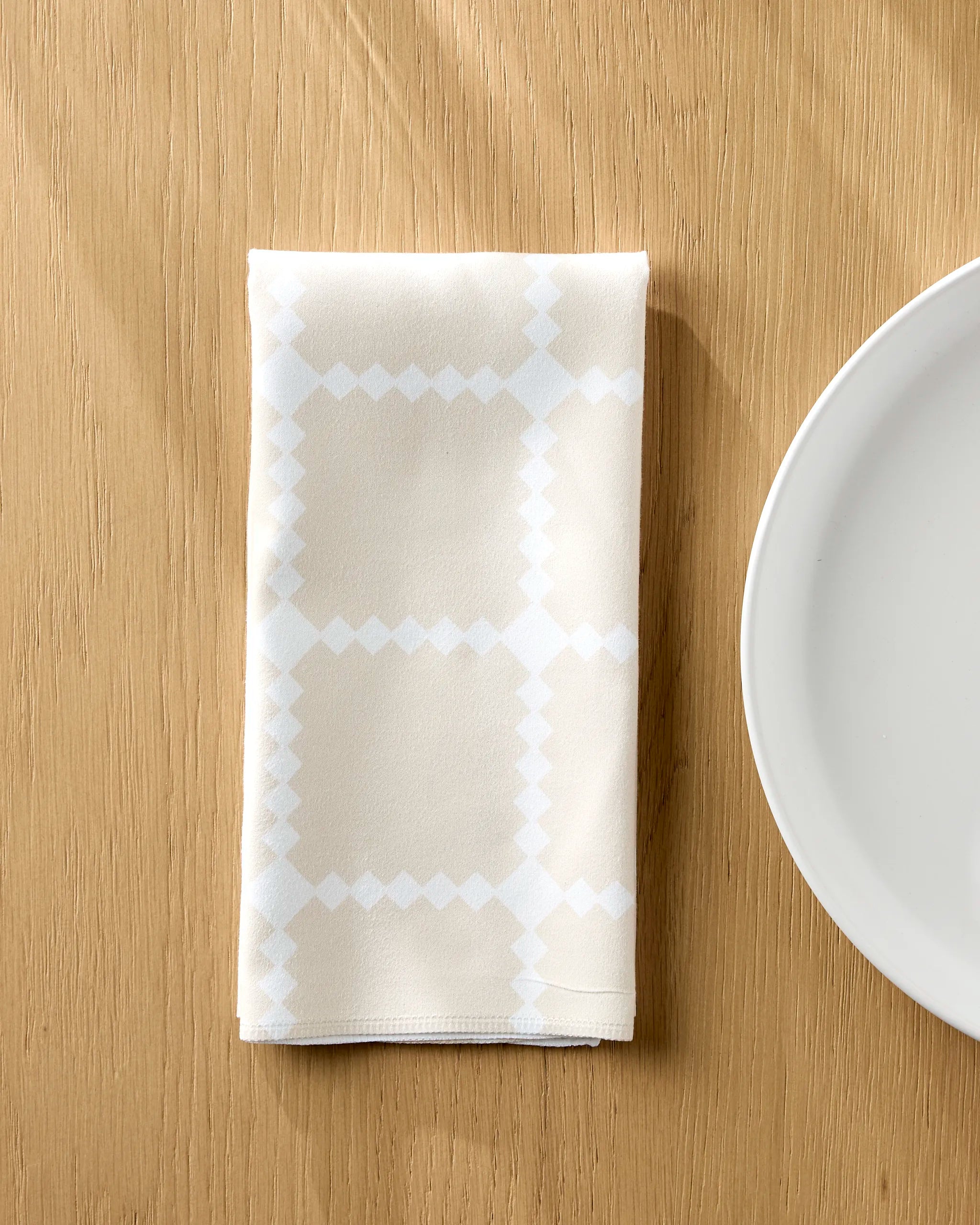 A folded white napkin from the Danelys Sidron Valencia Tile set, featuring a subtle geometric pattern, sits on a light wooden table next to a plain white plate.
