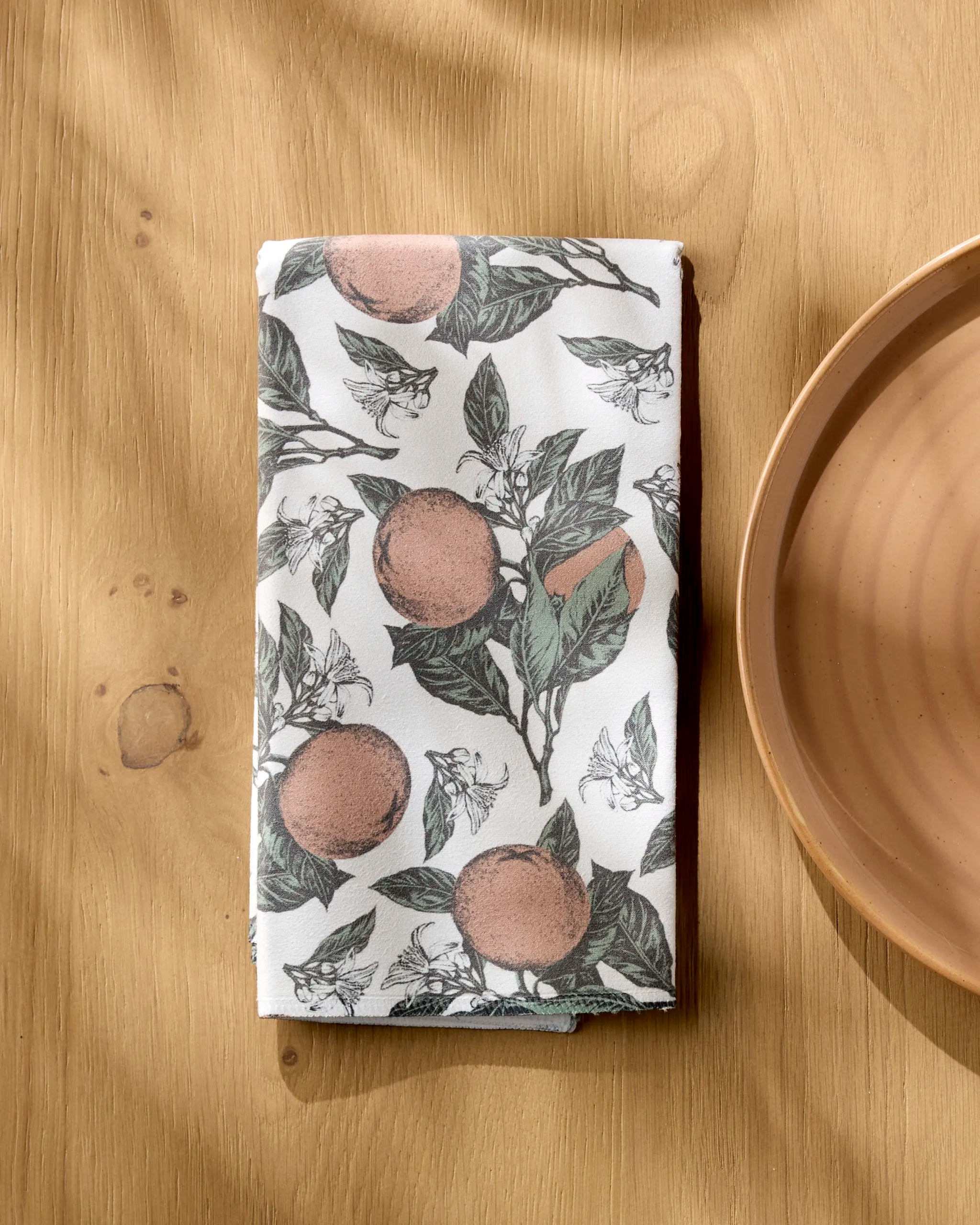 A folded white Valencia dinner napkin by Geometry, featuring an orange fruit and green leaf pattern, rests on a light wooden table beside a tan ceramic plate. Sunlight highlights this stylish and absorbent cloth napkin set.