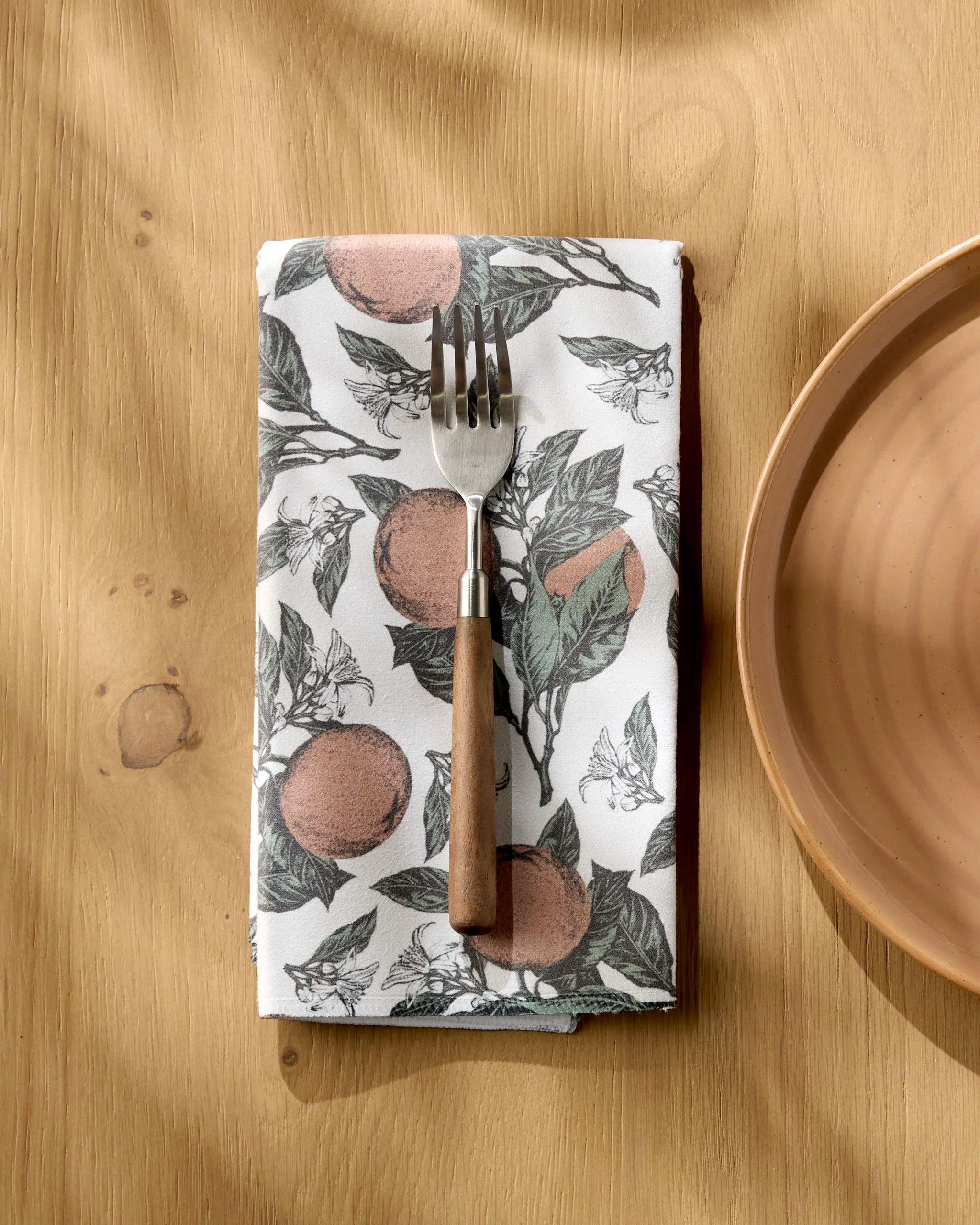 The Geometry Valencia fork with a wooden handle rests on an absorbent cloth napkin featuring an orange fruit and leaf pattern, beside a brown ceramic plate on a light wooden table.
