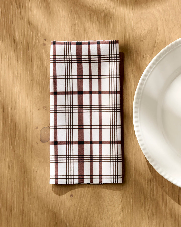 The Vintage Plaid Natural napkin by Hufton Studio, featuring brown and white checks, rests folded beside a scalloped white plate on a light wooden table. Sunlight casts gentle shadows over the scene.