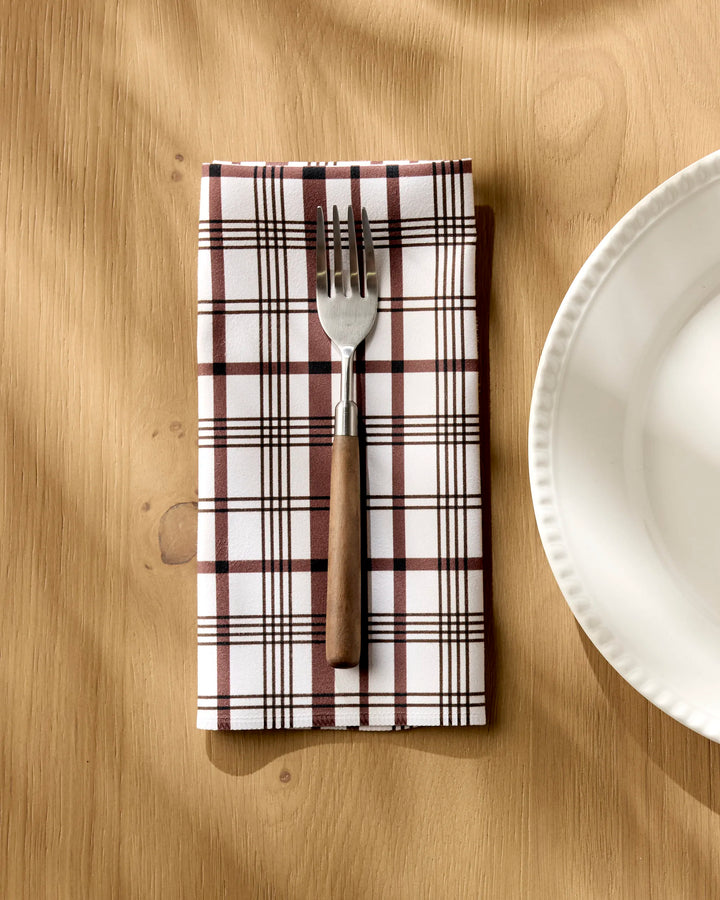 A fork rests on Hufton Studio’s Vintage Plaid Natural cloth napkin, placed on a light wooden table beside a white scalloped ceramic plate.