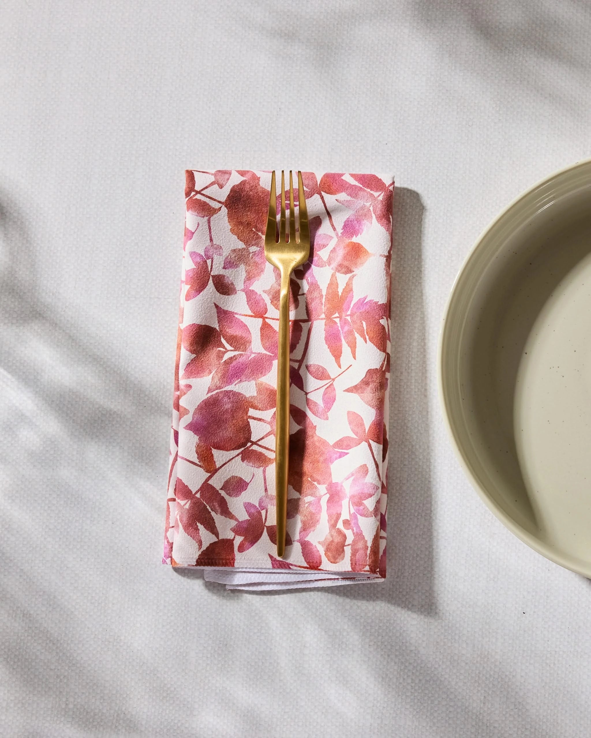 The Geometry Watercolor Dreaming eco-friendly napkin with a pink and red leaf pattern sits under a gold fork beside a beige plate on a white textured surface.