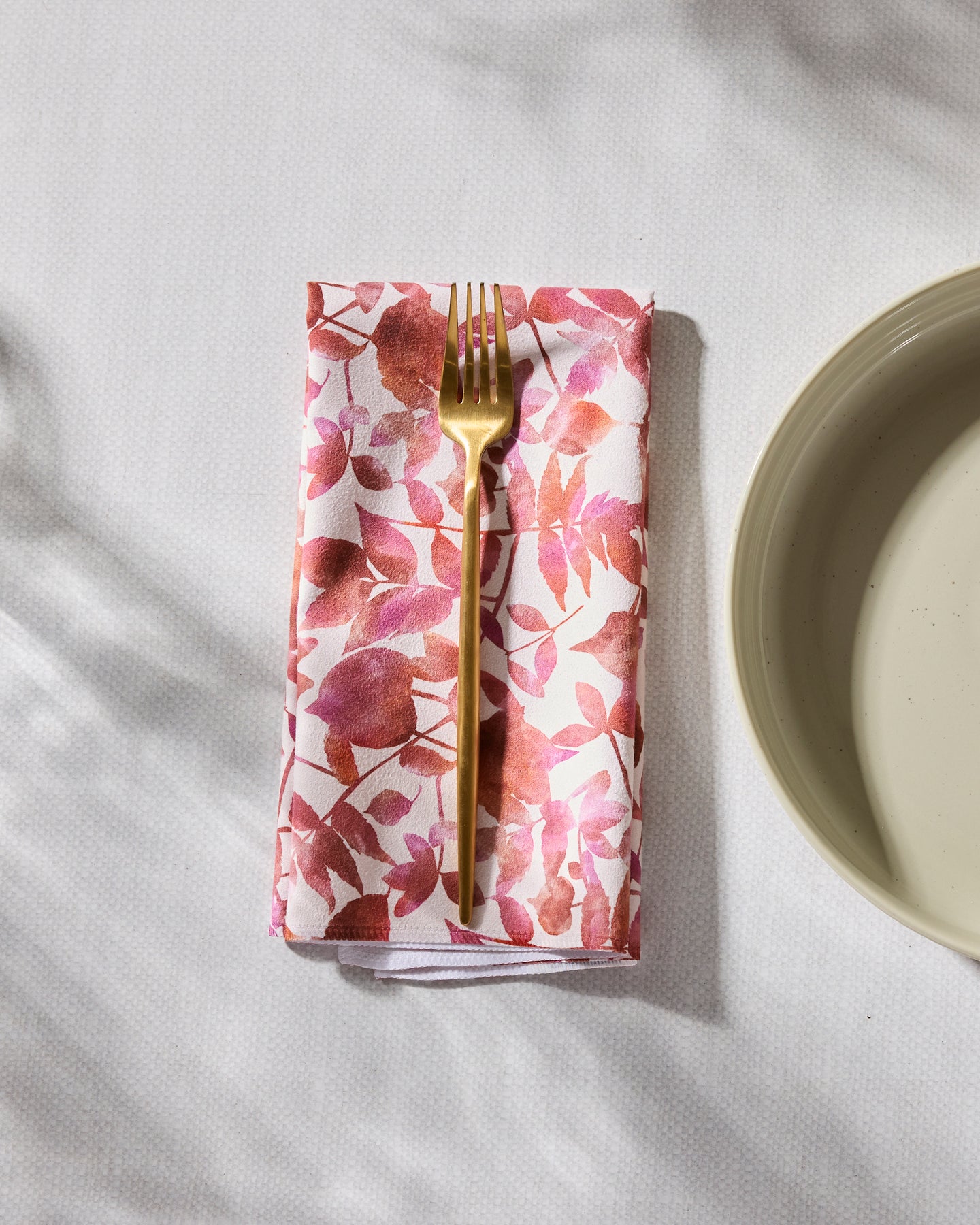 The Geometry Watercolor Dreaming eco-friendly napkin with a pink and red leaf pattern sits under a gold fork beside a beige plate on a white textured surface.