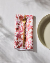 The Geometry Watercolor Dreaming eco-friendly napkin with a pink and red leaf pattern sits under a gold fork beside a beige plate on a white textured surface.