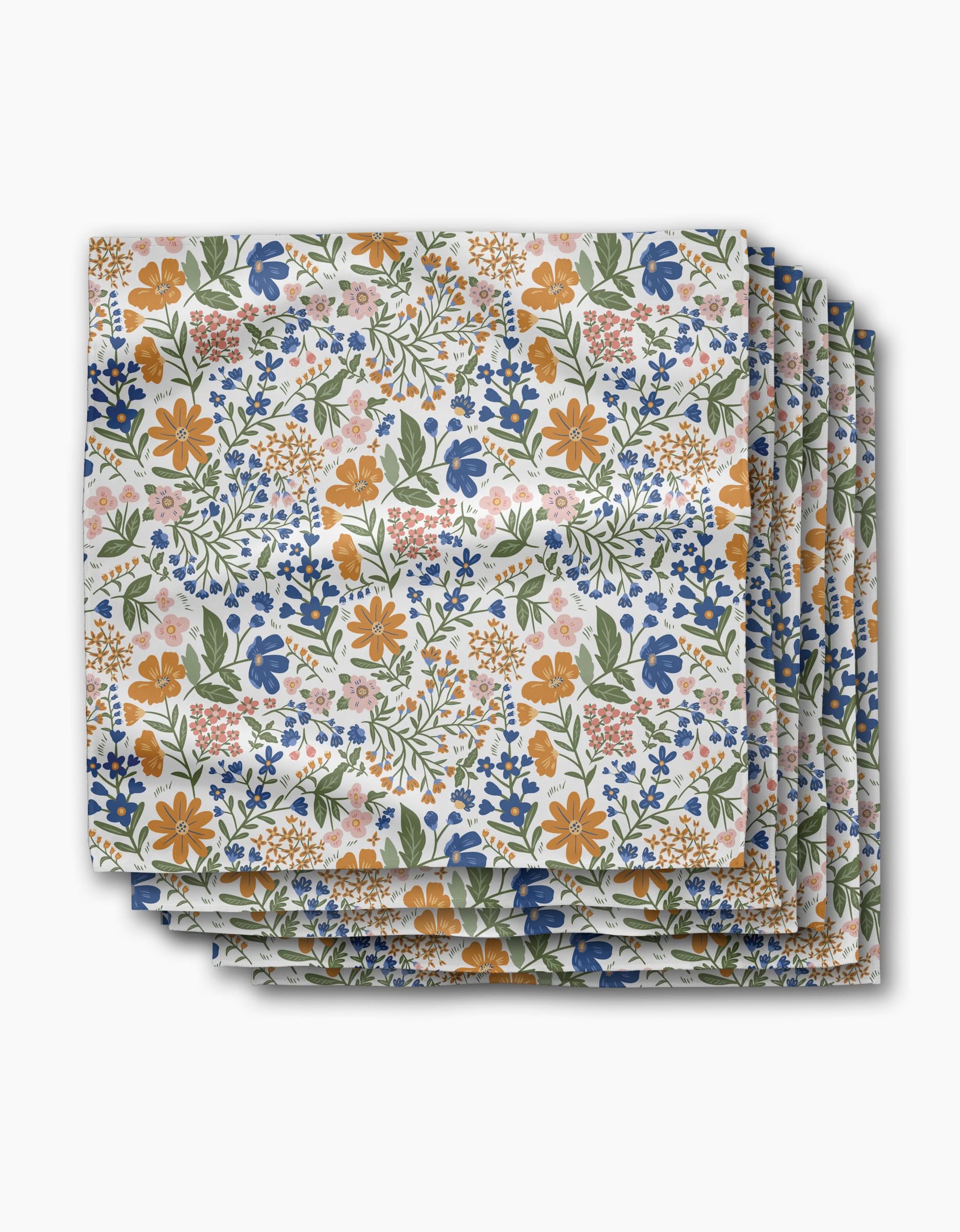 A stack of folded Danelys Sidron Wildflower Vine Blush Dinner Napkin Set, crafted from soft Geosuede with a vibrant floral pattern in orange, blue, pink, and green on white.