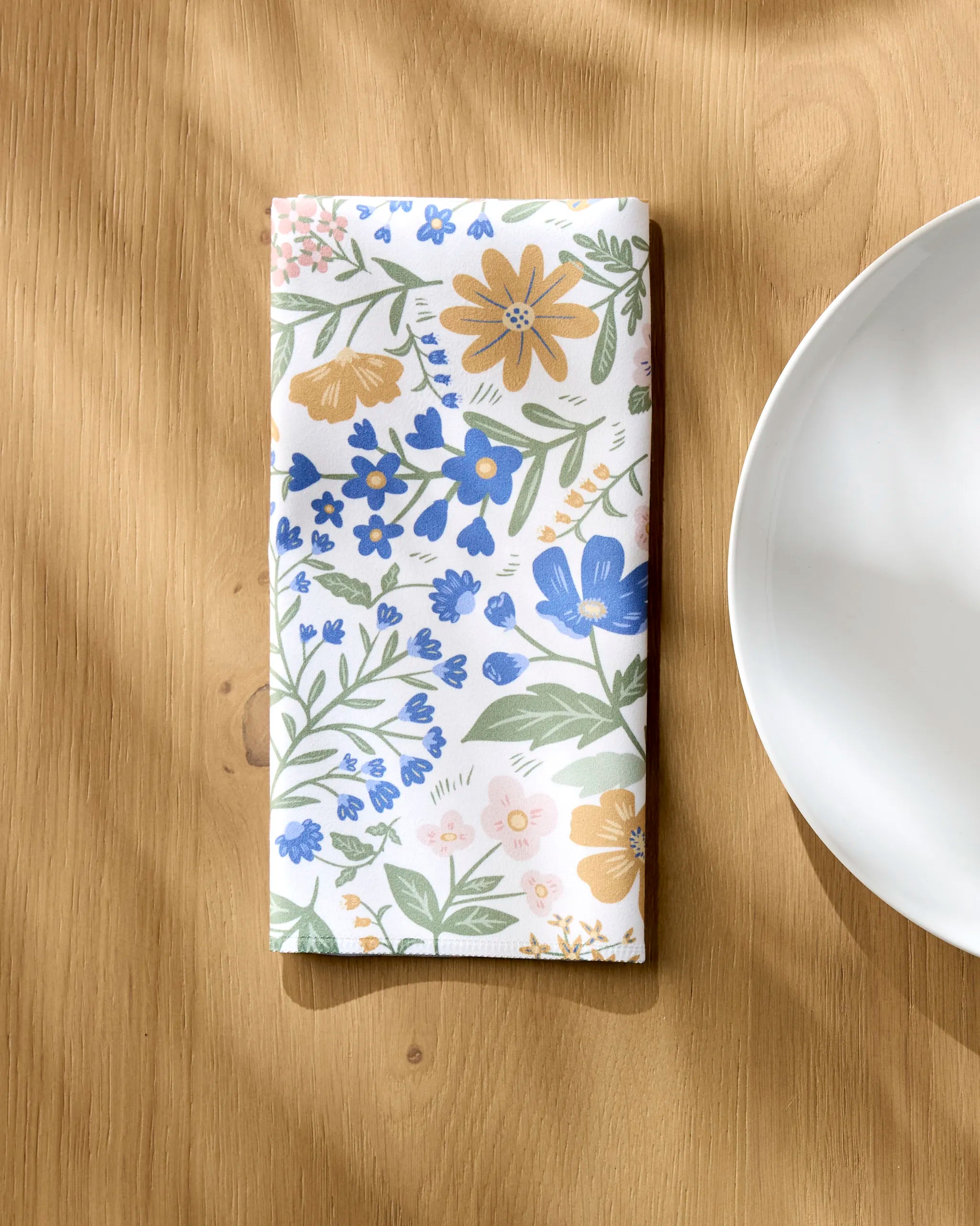 The Danelys Sidron Wildflower Vine Blush napkin set, featuring vibrant florals on Geosuede fabric, is neatly folded on a wooden table beside a white plate. Sunlight highlights the elegant, recycled design.