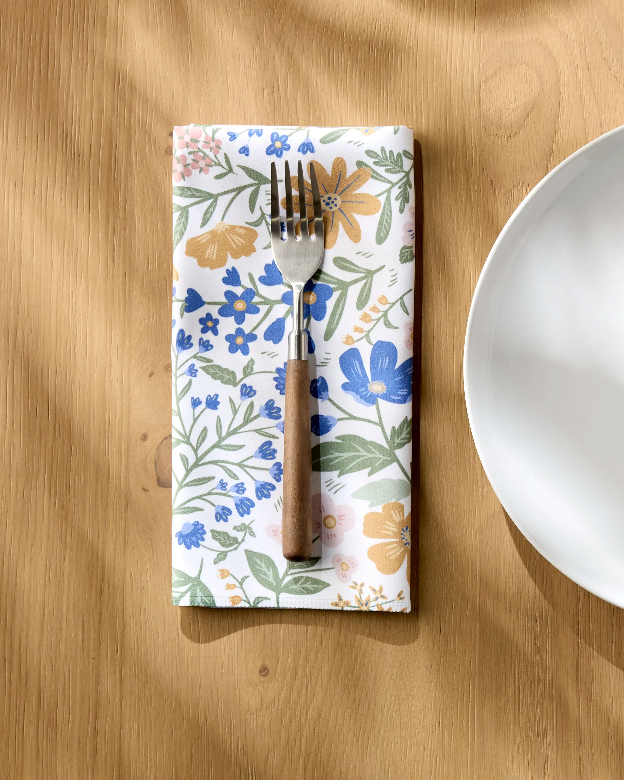 A fork with a wooden handle rests on the Danelys Sidron Wildflower Vine Blush napkin, set on a light wood table beside a white plate to the right. Sunlight casts soft shadows, highlighting this elegant dinner setting.