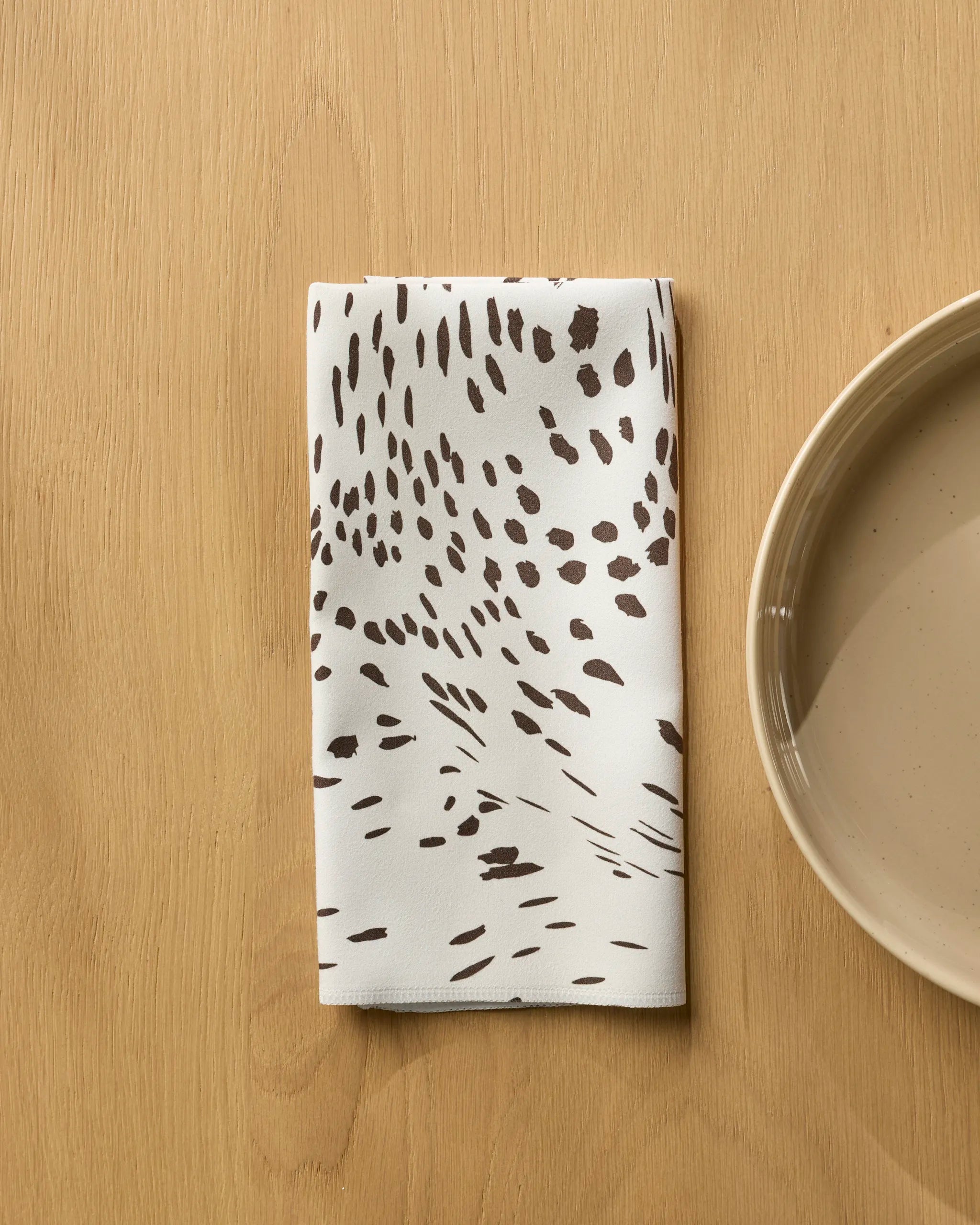 The Geometry Wildgrain folded white reusable napkin, featuring an abstract brown dotted pattern, rests on a light wooden table beside a beige ceramic plate.
