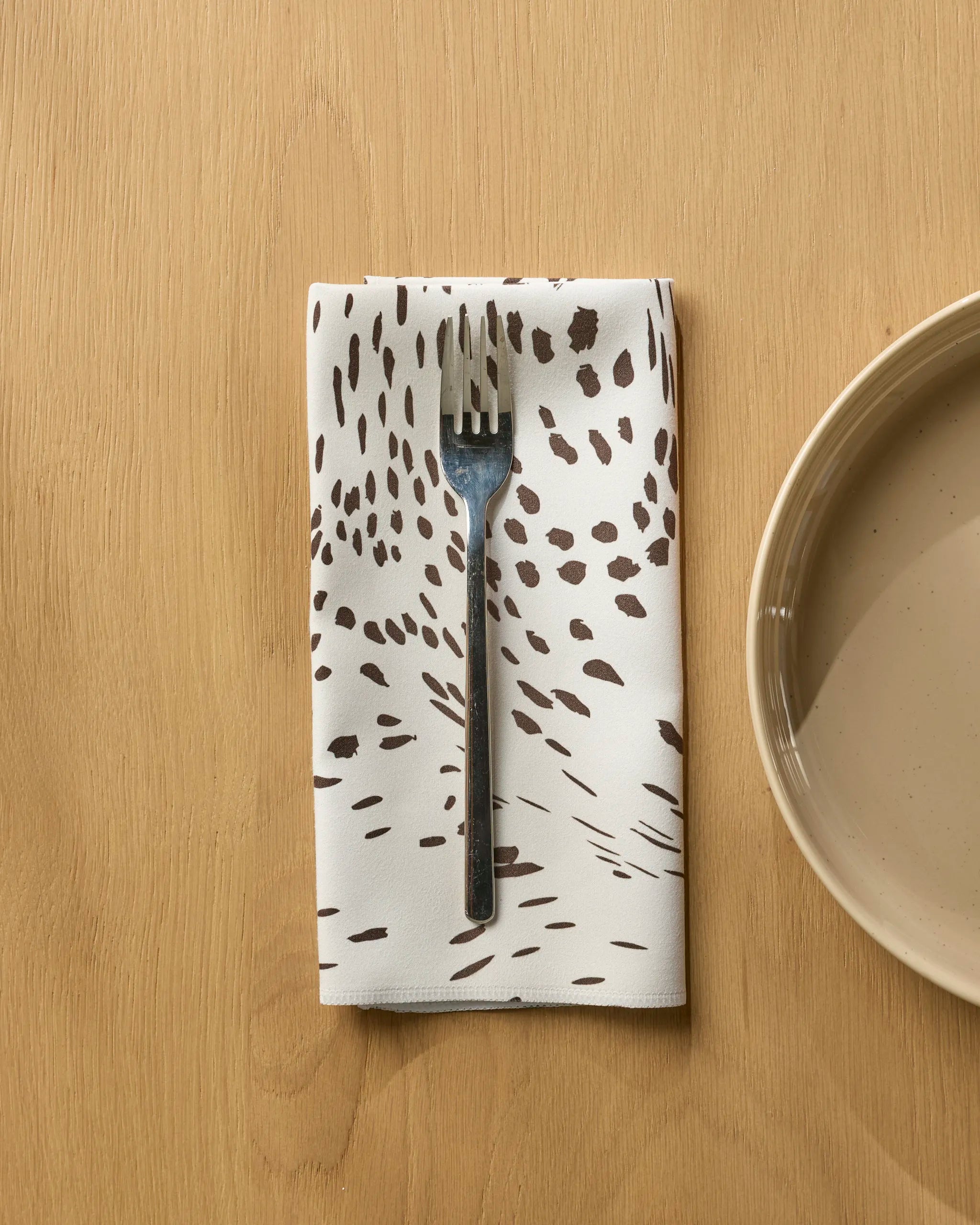 A silver fork rests on a folded Wildgrain reusable napkin by Geometry, featuring brown abstract spots, set on a light wooden table beside a beige ceramic plate.
