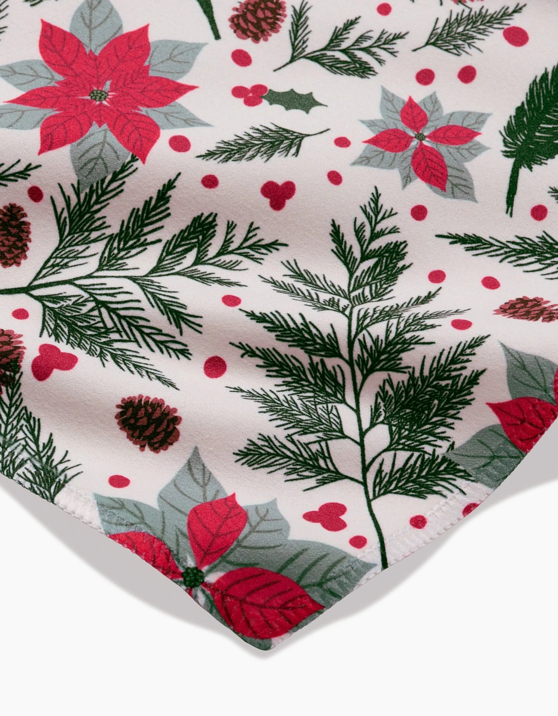 A close-up of Rebecca Bobkos Woodland Poinsettia Geosuede fabric displays red poinsettias, green pine branches, pinecones, holly leaves, and red berries on white—ideal for recycled dinner napkins.