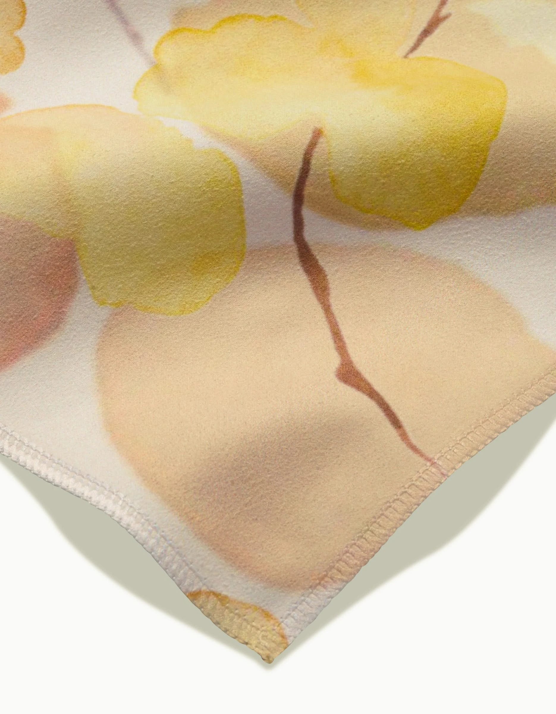 Close-up of the GEOMETRY Yellow Fall dinner napkin in soft Geosuede fabric, featuring a watercolor floral pattern in light yellow and peach with brown stems and stitched edging.
