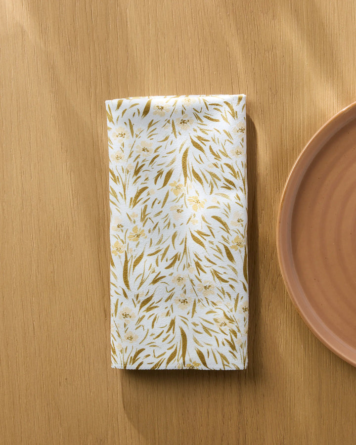 A neatly folded Melissa Johnson Yellow Floral reusable dinner napkin rests on a light wooden table beside a round, light brown ceramic plate, with sunlight casting soft shadows across the scene.