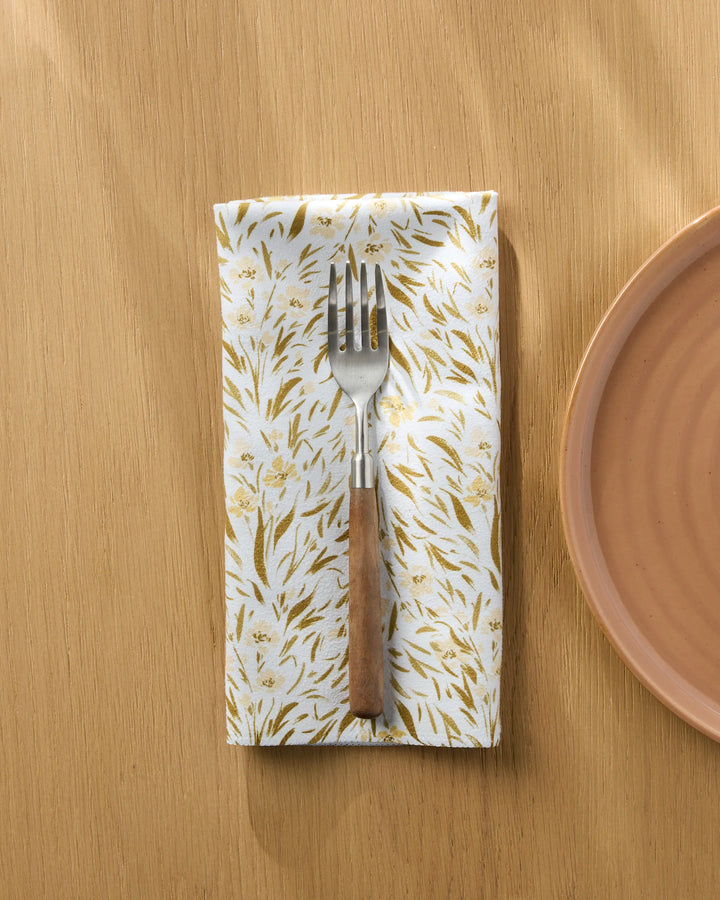 A Melissa Johnson Yellow Floral reusable dinner napkin holds a fork with a wooden handle, resting beside the edge of a round, light brown plate on a light wooden table.