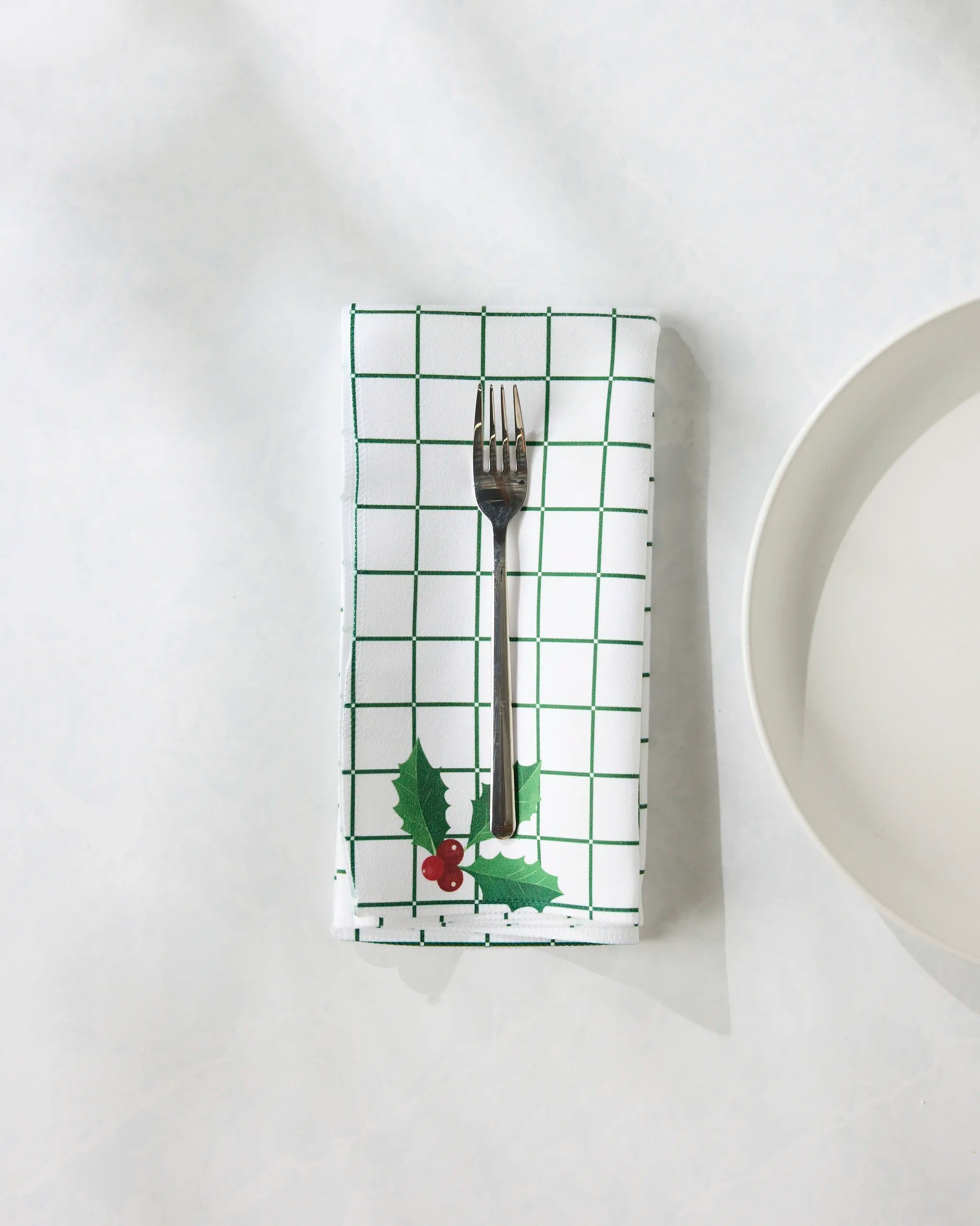 A metal fork rests on Geometry’s Yuletide Grid eco-friendly napkin, featuring a green grid and holly design, beside a white plate on a light surface.