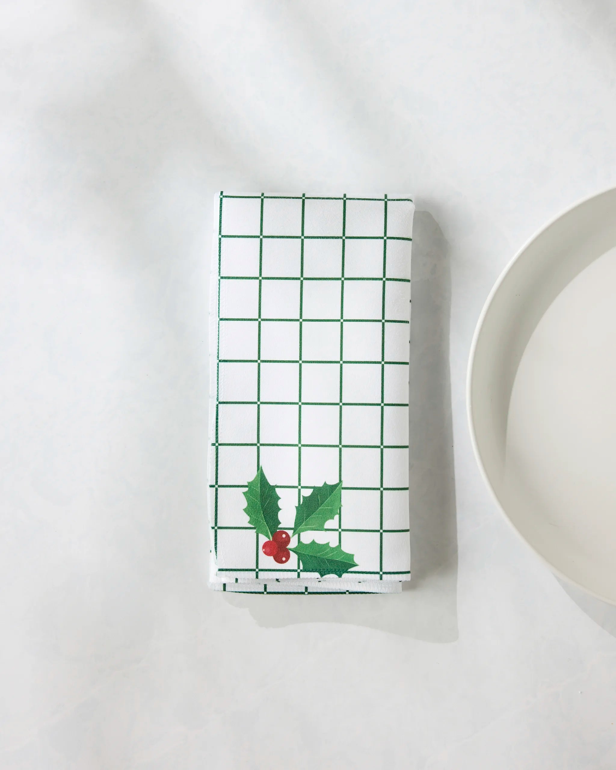 The Geometry Yuletide Grid napkin, featuring an eco-friendly white fabric with a green grid and holly leaf design, is folded neatly beside a white plate on a light surface.
