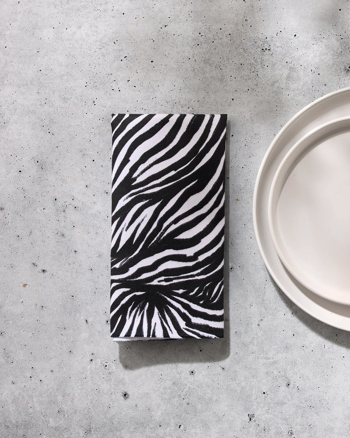 The GEOMETRY Zebra reusable cloth napkin with a black and white zebra print is folded on a light gray speckled surface beside a white plate.