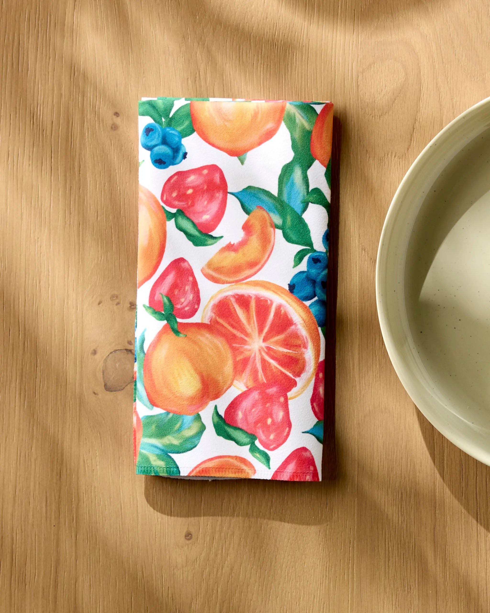 A folded Aubrey Fairchild Zesty Life napkin with a vibrant fruit pattern rests on a wooden table beside a cream ceramic bowl, sunlight casting soft shadows across the scene.