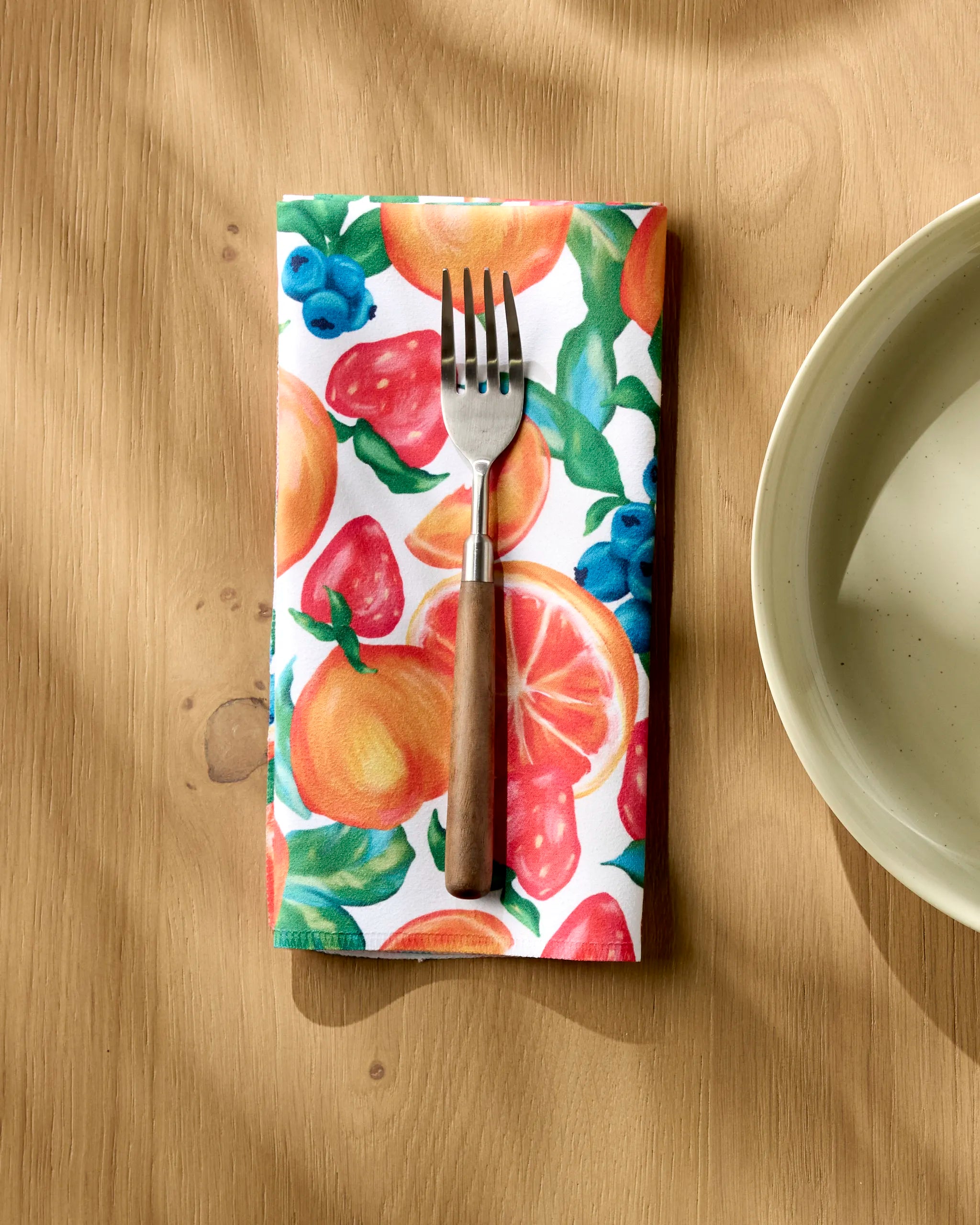 A fork with a wooden handle rests on Aubrey Fairchild’s Zesty Life absorbent cloth napkin, featuring a colorful fruit pattern, beside a beige plate on a light wood table lit by sunlight.