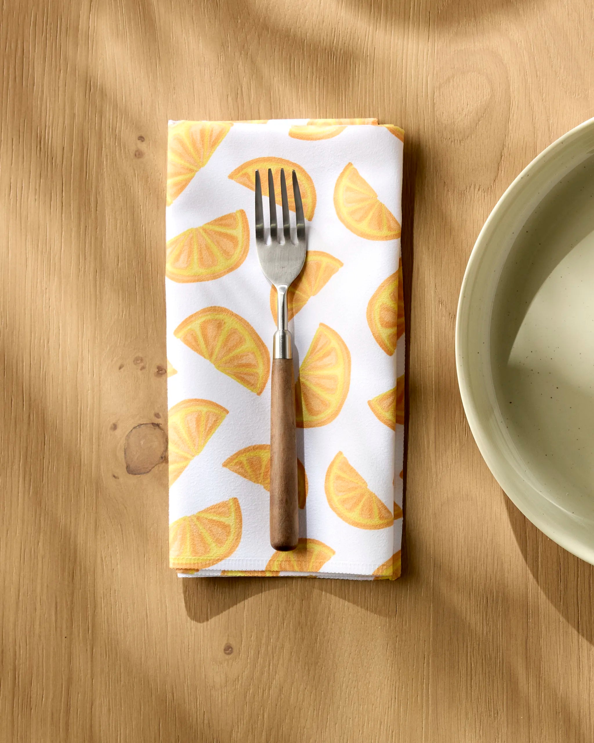 The Aubrey Fairchild Zesty Slice fork with a wooden handle rests on an absorbent, lemon-patterned napkin atop a light wood table beside a cream ceramic bowl, as sunlight casts gentle shadows over the scene.