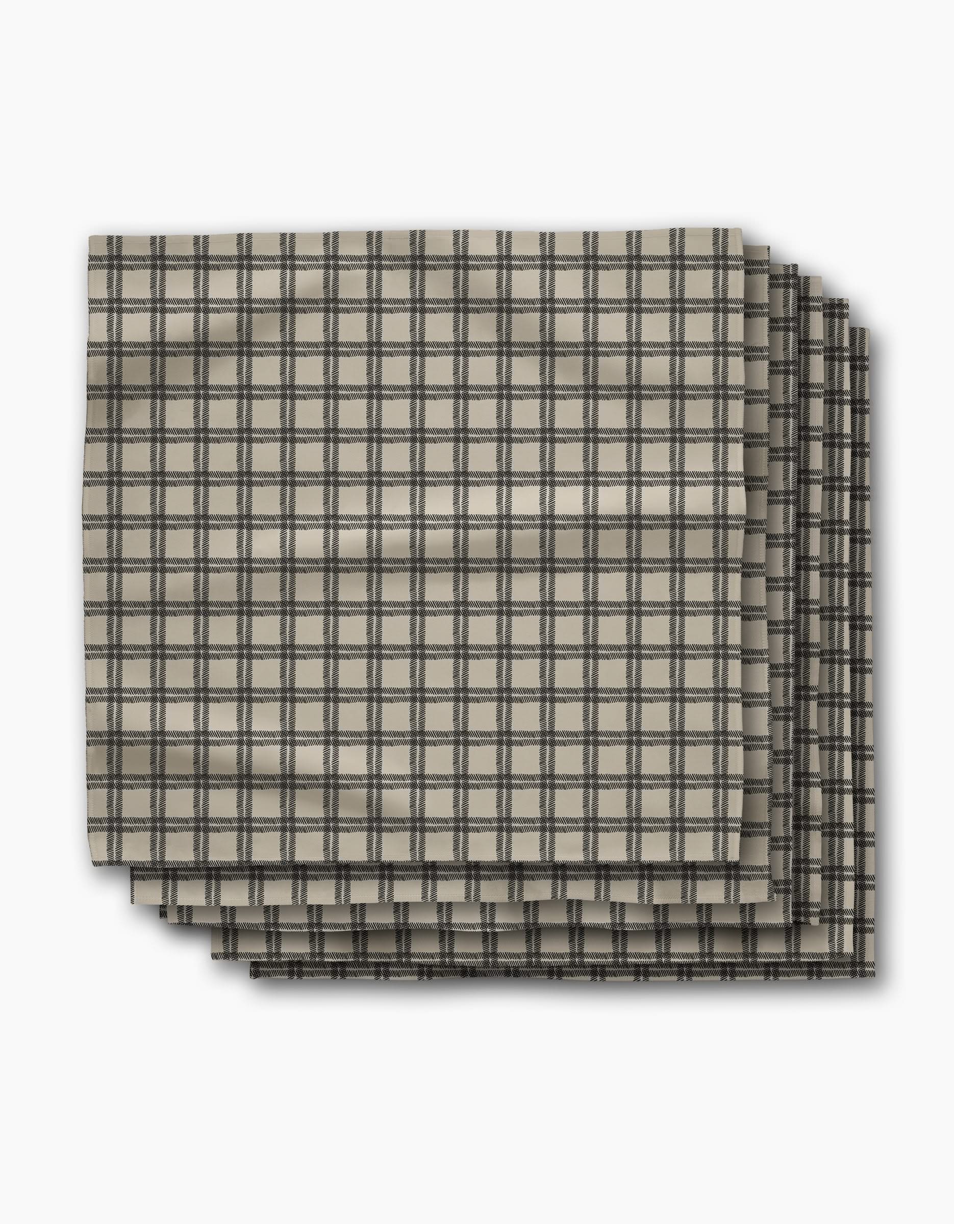 Four beige fabric napkins with a black grid plaid pattern, the Blackstone Dinner Napkin Set by Geometry, are neatly stacked. Crafted from absorbent recycled materials, this set is both stylish and practical.