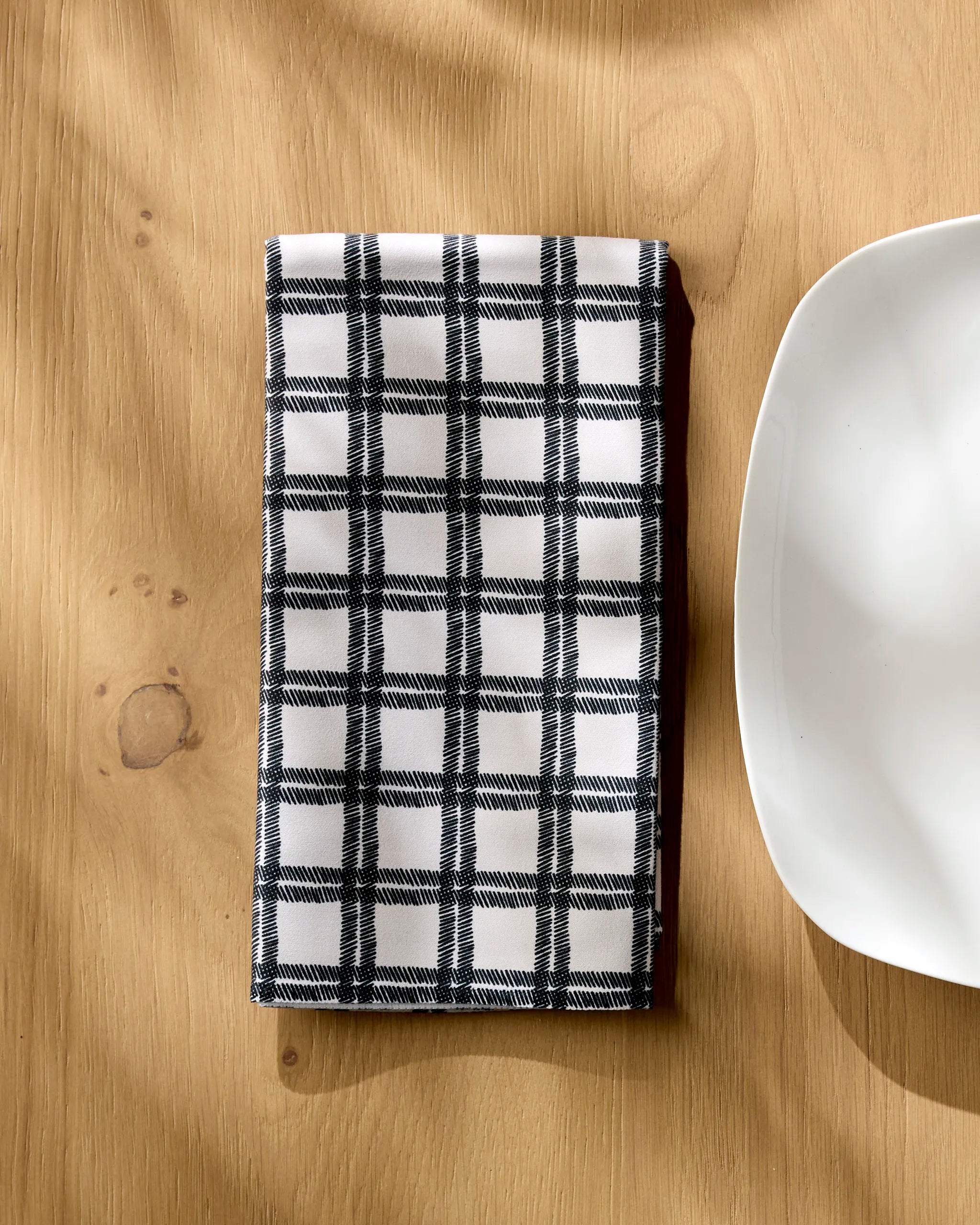 A Geometry Blackstone recycled napkin with a black and white checkered pattern is neatly folded on a light wooden table beside the edge of a white, square plate.