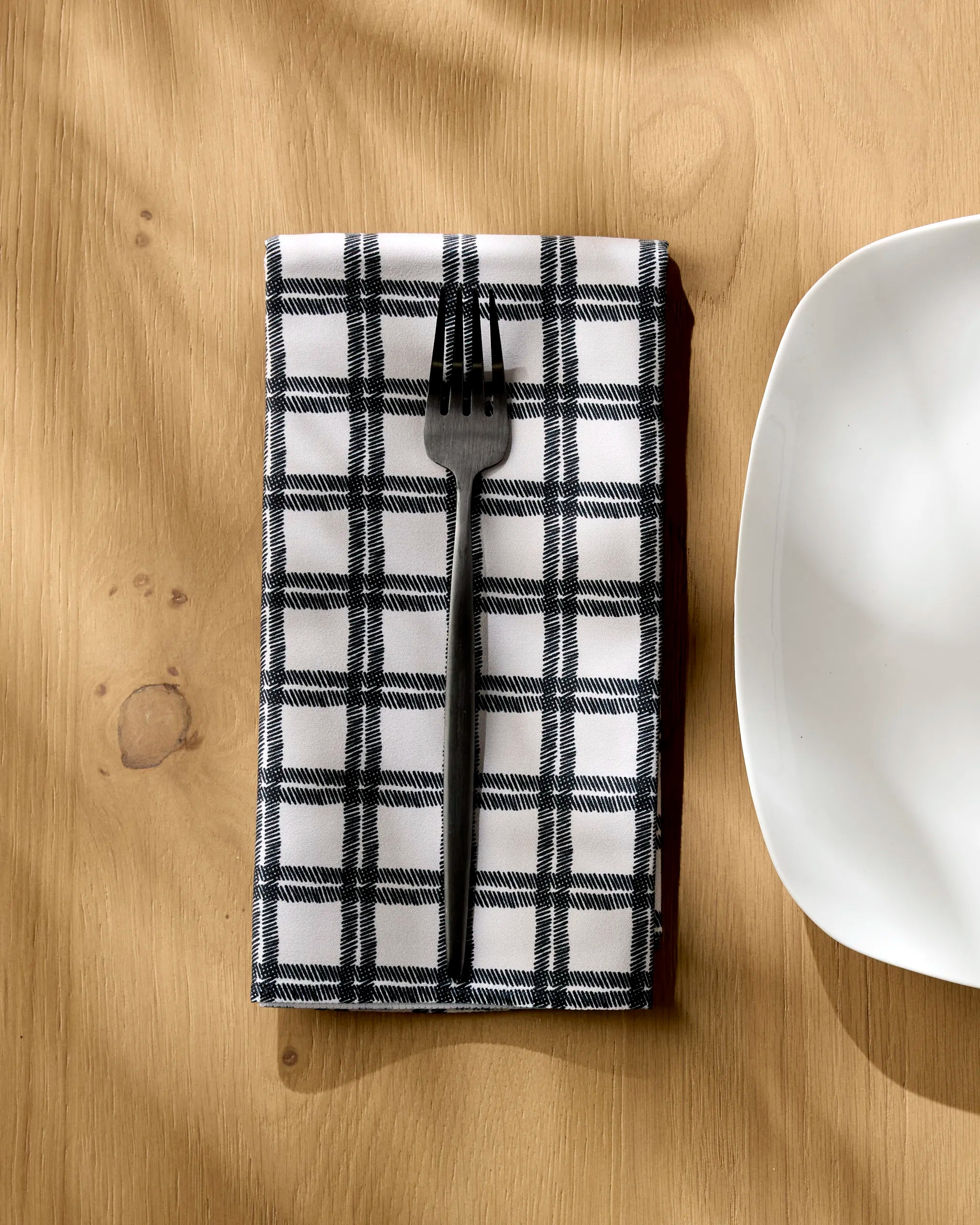 A Blackstone fork by Geometry rests on a folded, black and white checkered recycled napkin on a wooden table beside a rounded-edge white plate.