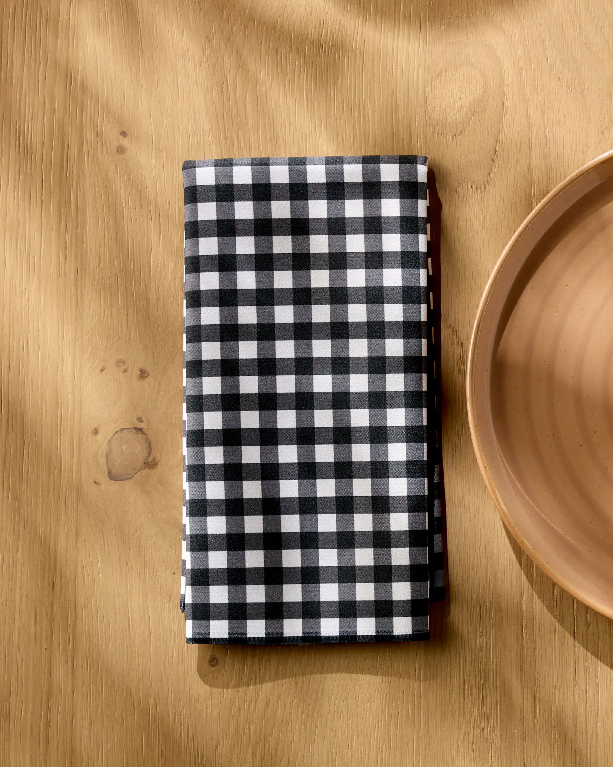 The Geometry Charcoal Gingham eco-friendly napkin, neatly folded, rests on a wooden table beside a plain light brown ceramic plate.