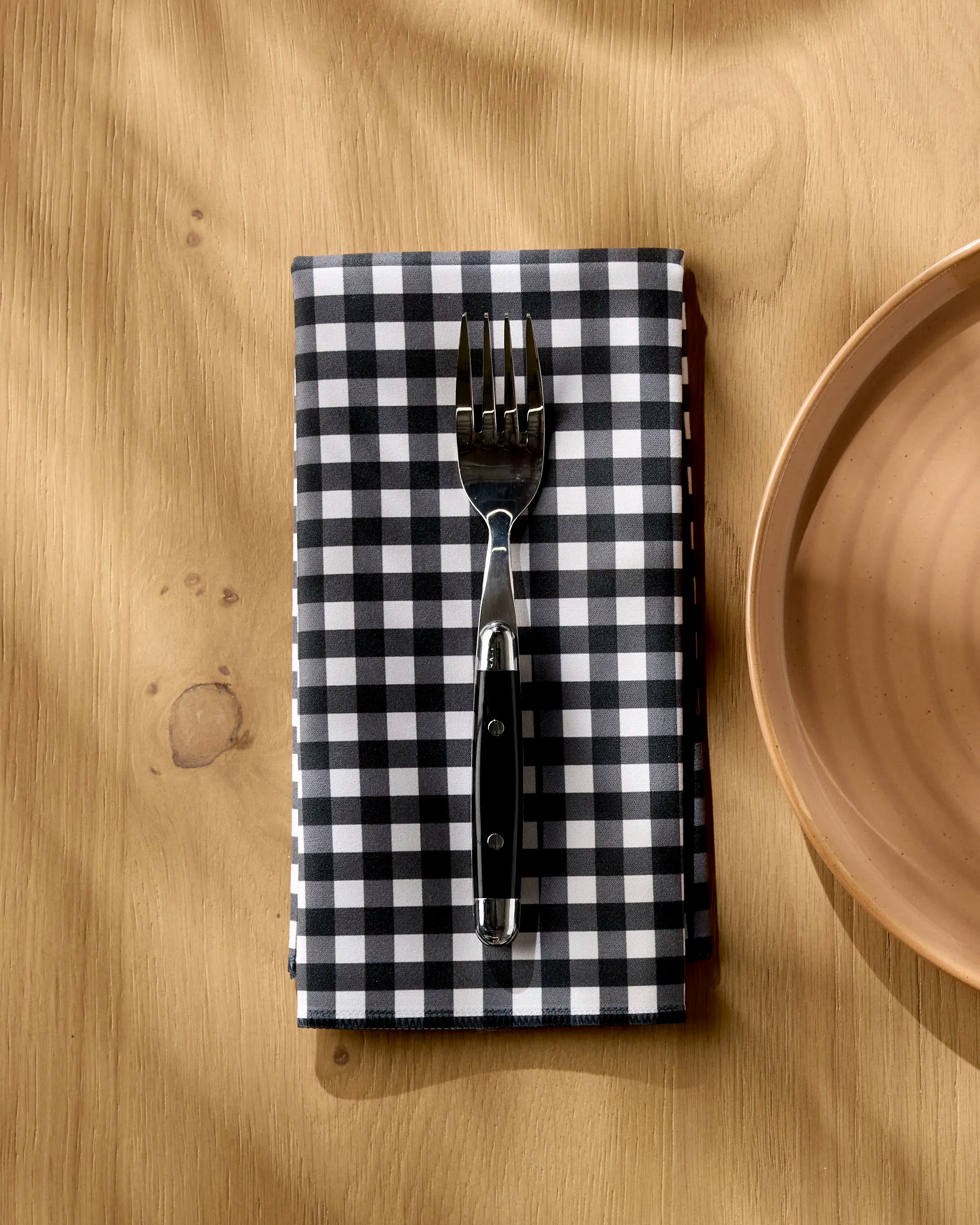 A fork rests on an absorbent Geometry Charcoal Gingham napkin, featuring a black-and-white checkered pattern, on a light wooden table next to an empty beige ceramic plate.