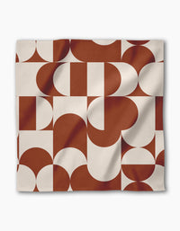 The Circle Playground napkin by Geometry is a square, reusable cloth with an abstract geometric pattern in rust brown and off-white, featuring curved and straight shapes for a modern look. The fabric displays soft folds and subtle shadows.