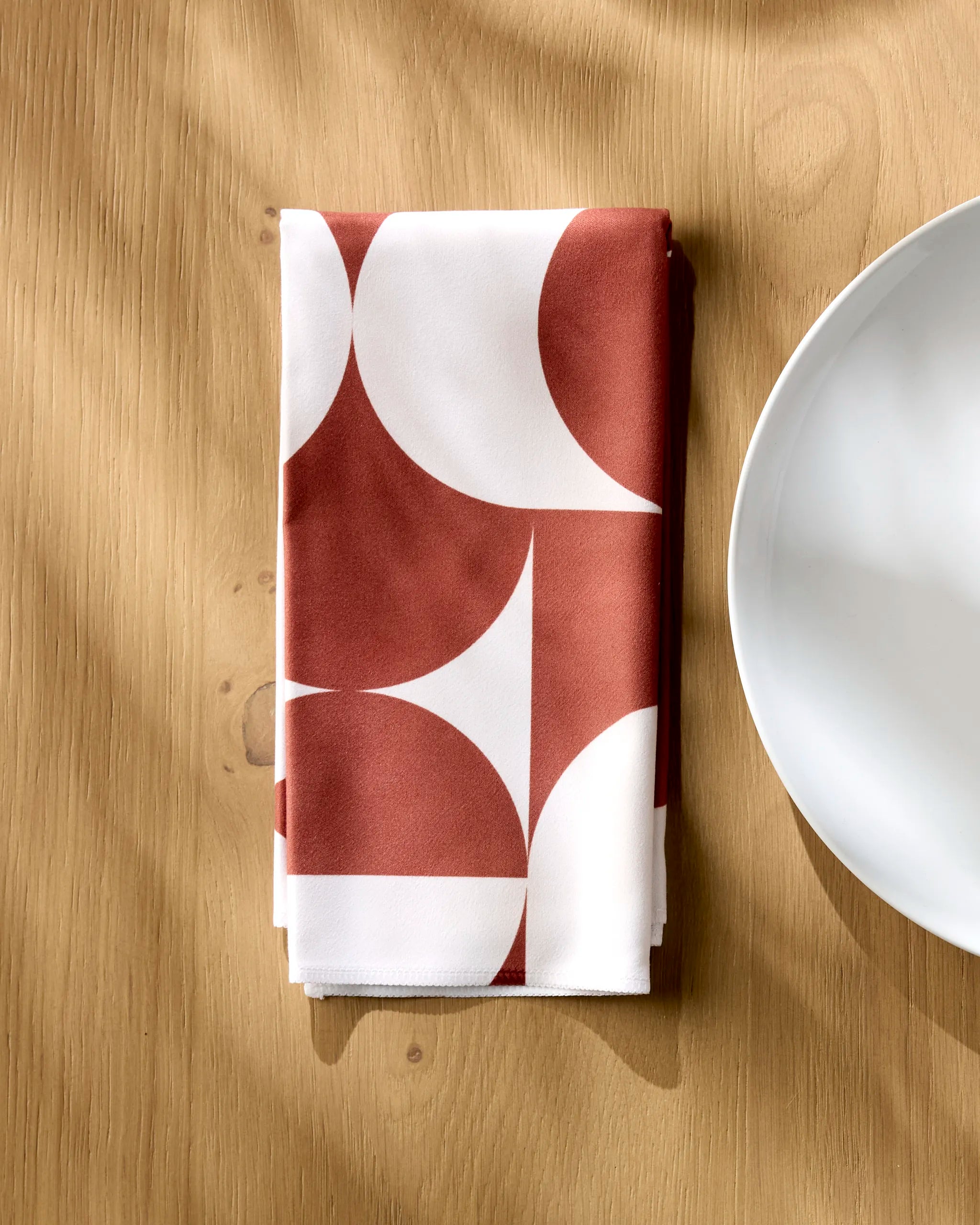 The Geometry Circle Playground reusable napkin, featuring a bold red and white geometric pattern, rests on a light wooden table beside a plain white plate as sunlight casts gentle shadows across the scene.