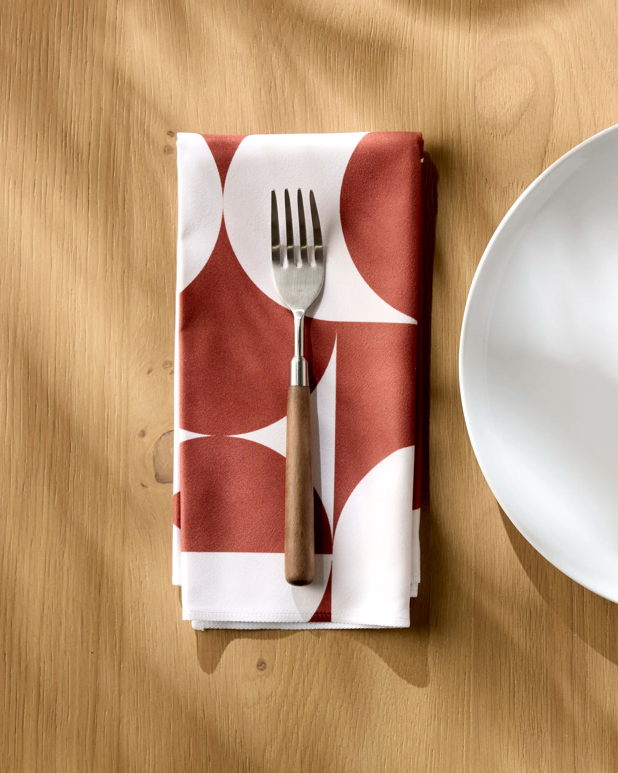 The Circle Playground reusable napkin by Geometry, featuring a red and white geometric design, is folded on a light wooden table with a fork placed on top beside a white plate.